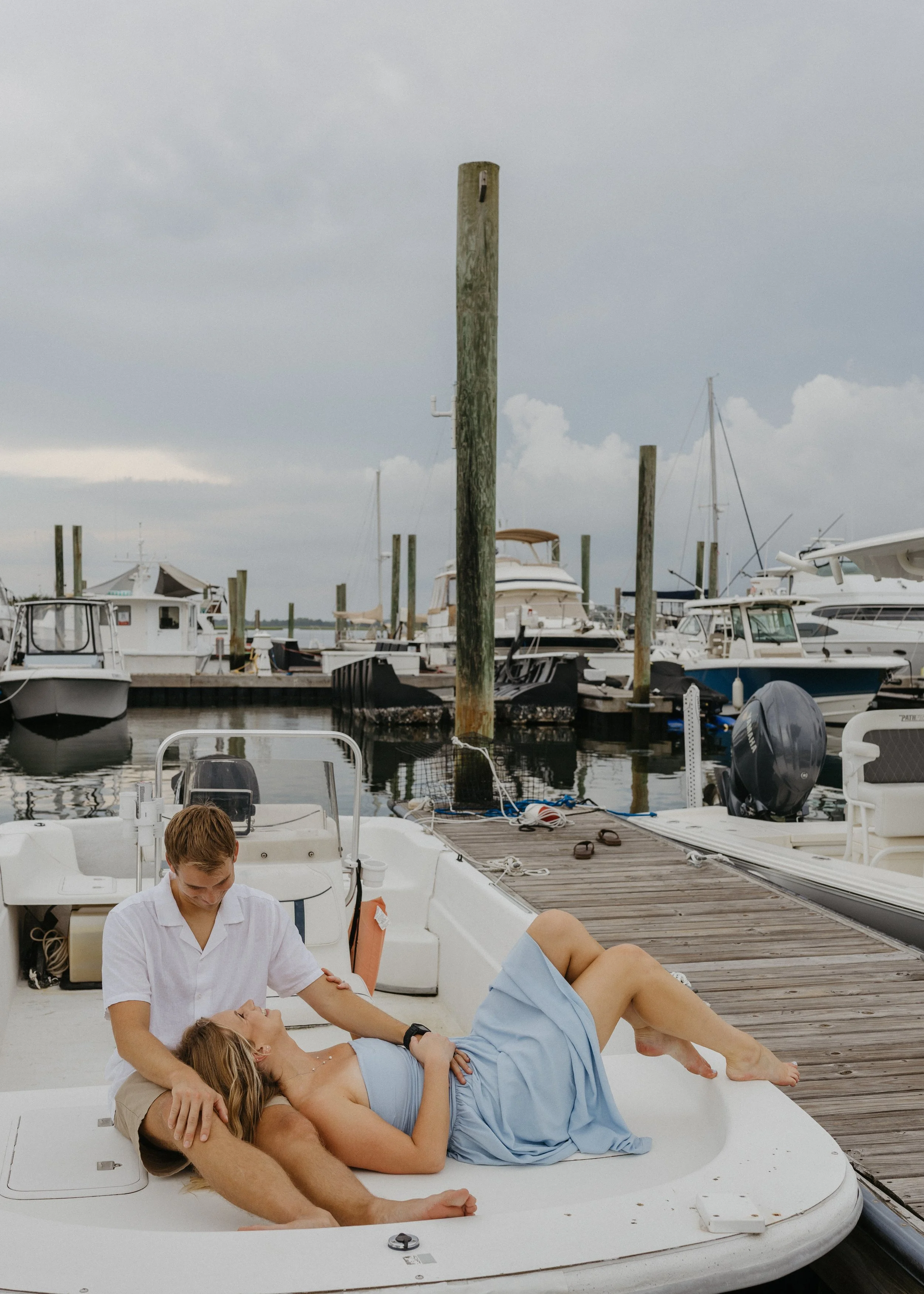 A couple laying on the deck of a boat with a man in a white shirt beside them, on a dock at a marina with several boats and boats on land in the background.