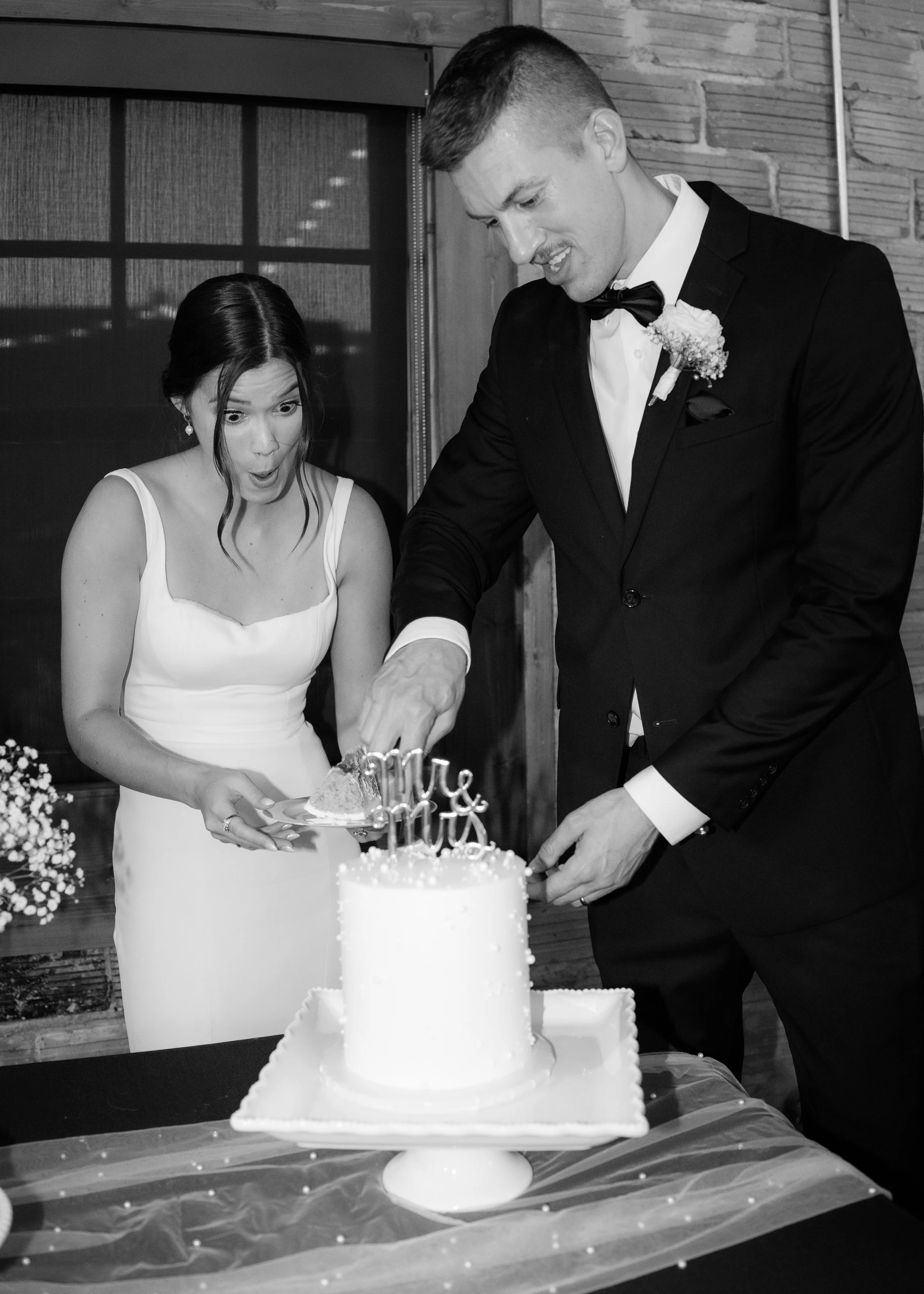 Black and white photo of a bride and groom cutting a wedding cake together at their wedding reception. The bride has dark hair styled in an elegant updo with loose curls and is wearing a sleeveless wedding dress. The groom is dressed in a tuxedo with