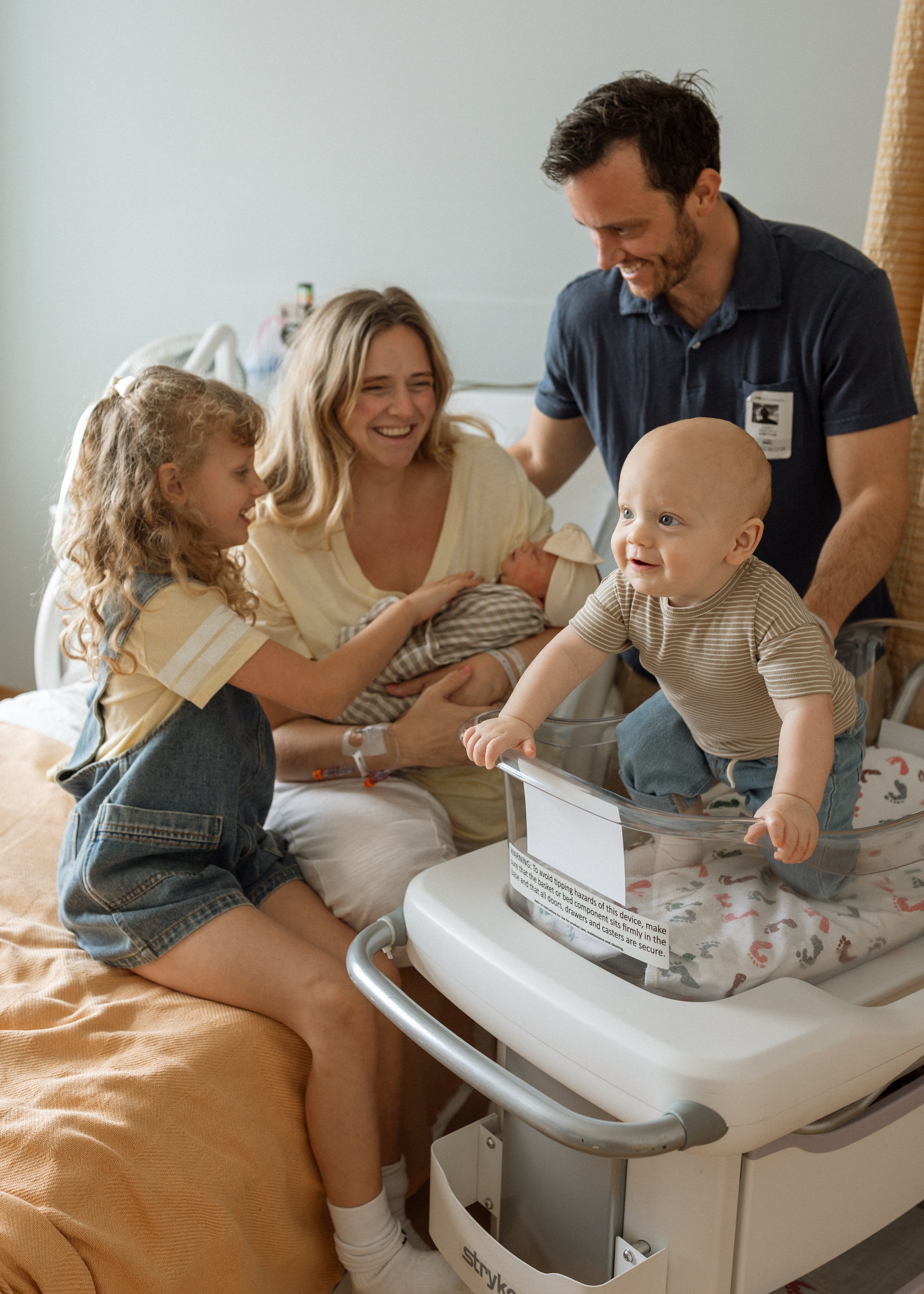 A woman holding a newborn baby in a hospital bed, with a young girl and a man smiling at a baby in a hospital bassinet.