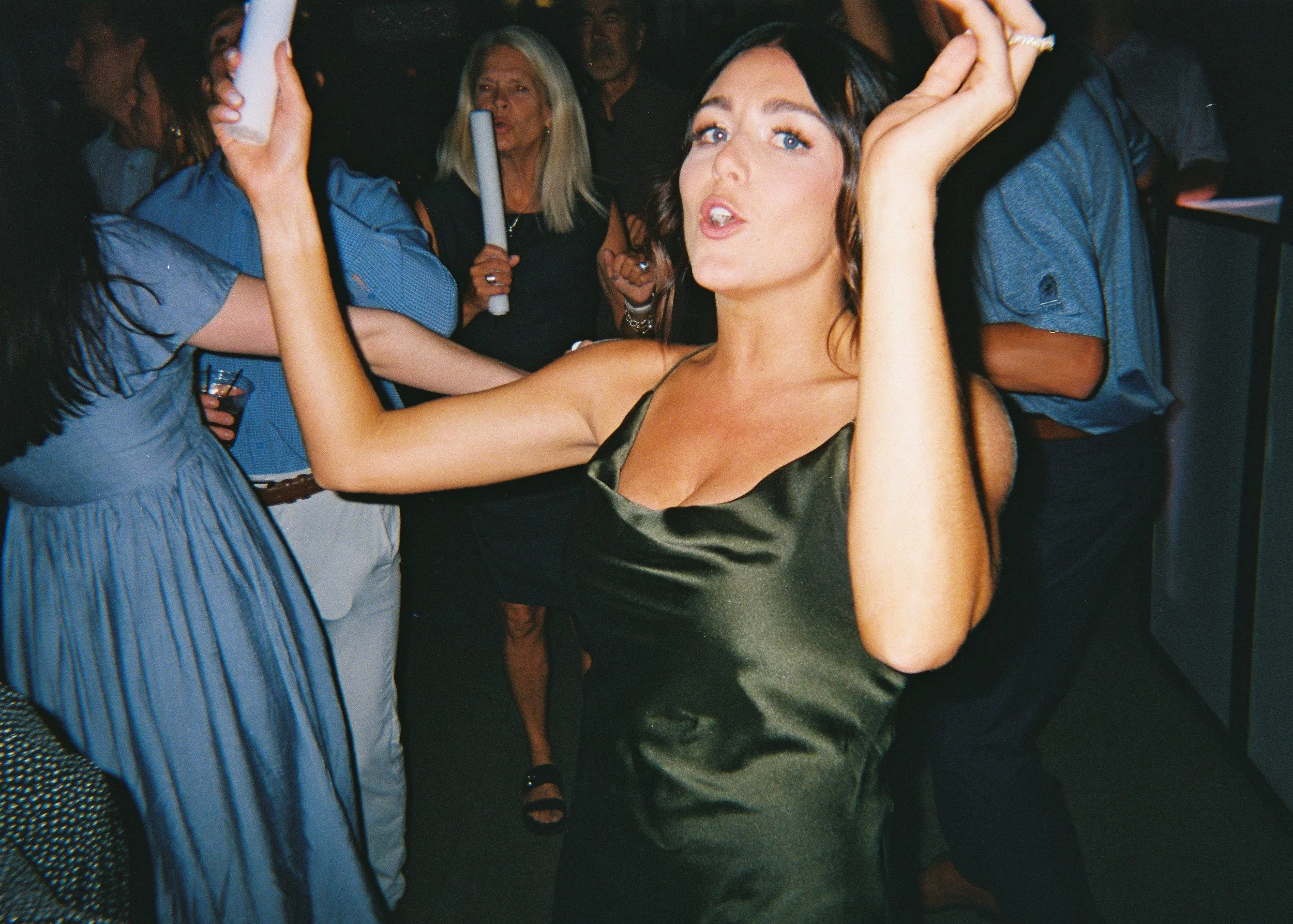 A young woman with black hair and blue eyes taking a selfie in a crowded event. She is wearing a dark satin dress, with her right arm raised holding a phone. People are visible in the background, some holding drinks or props.