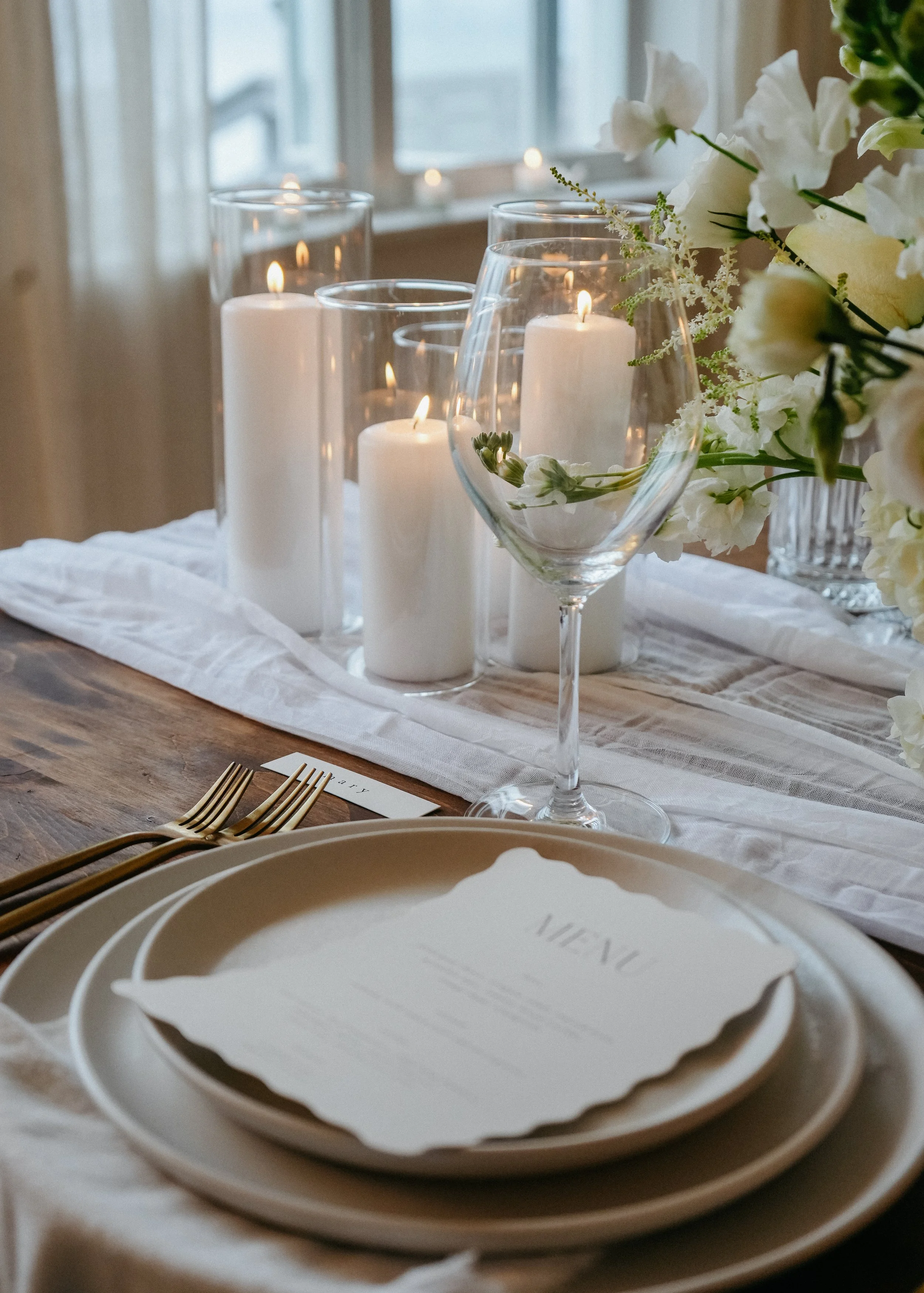 Rustic dining table decorated with lit white candles in glass holders, a wine glass with flowers, and a floral arrangement, set near a window with soft natural light.