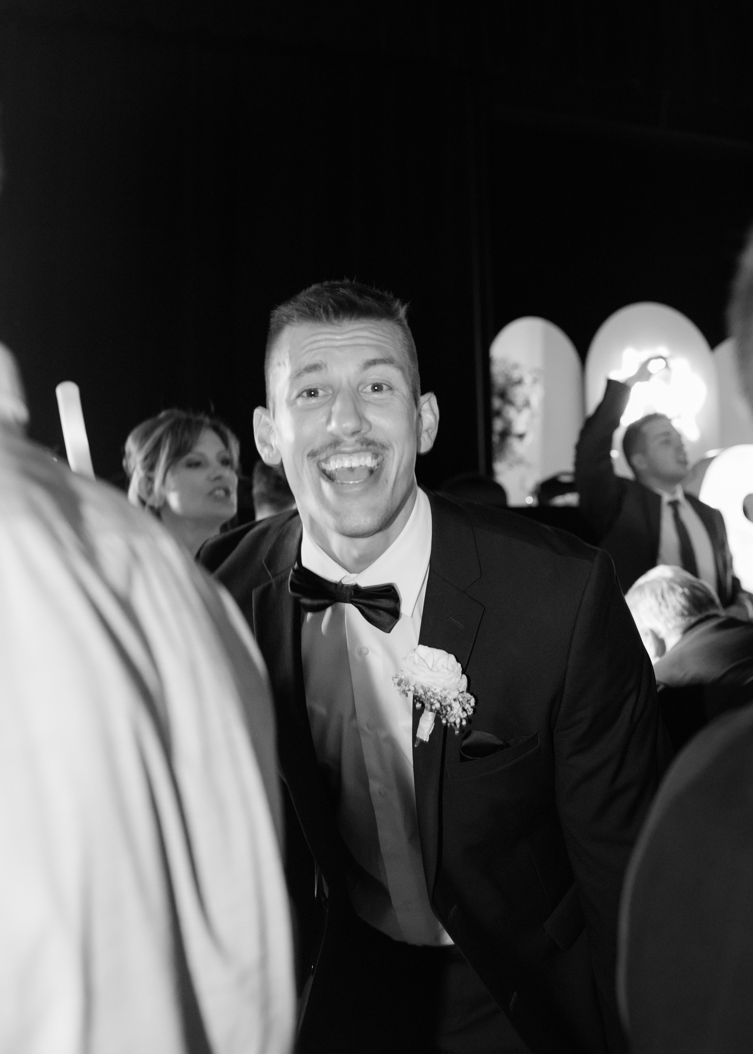 Man wearing a tuxedo and bow tie, smiling, at a formal event with other people in the background.