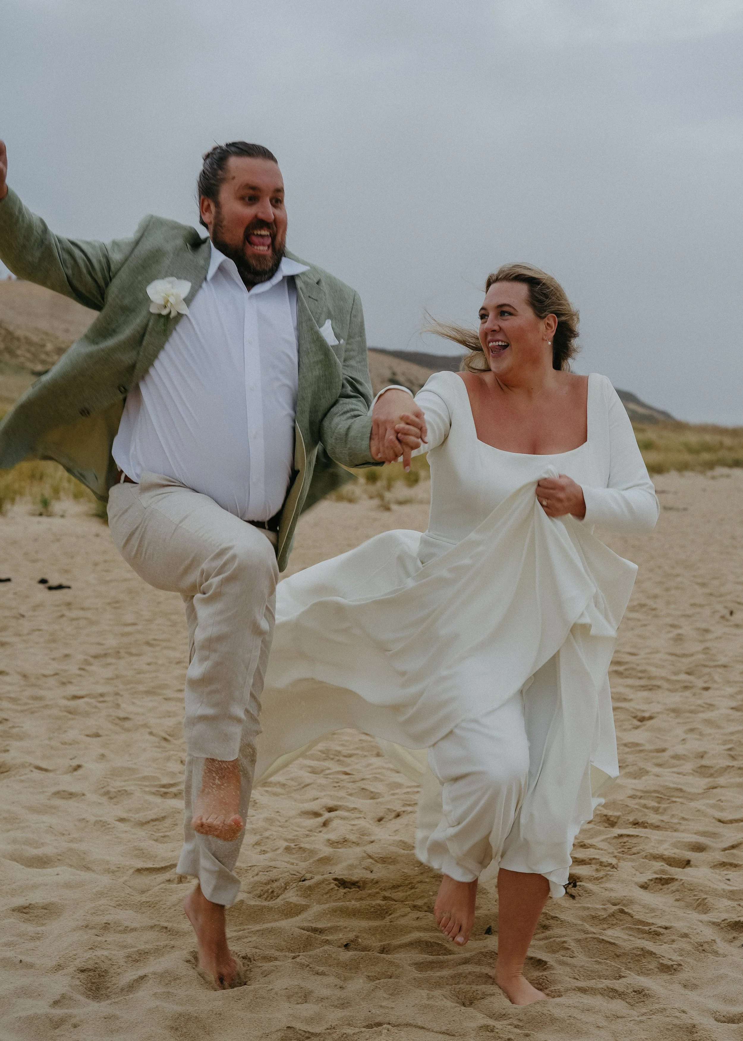 A man and woman in wedding attire running barefoot on a sandy beach, smiling and holding hands, with a cloudy sky and hills in the background.