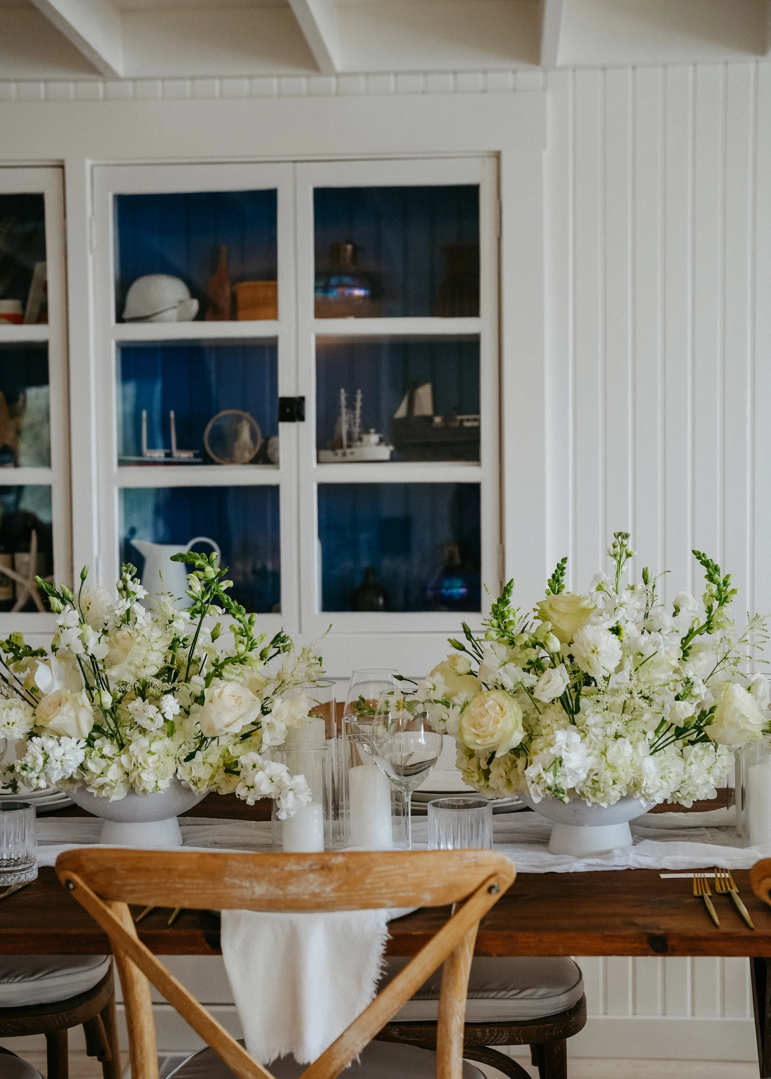 A dining table decorated with white flower arrangements, candles, and glassware, with a white-paneled wall and a cabinet with glass doors in the background.