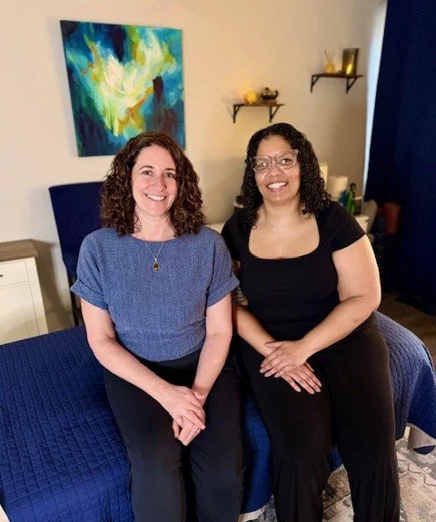 Two women sitting on a bed in a bedroom with colorful artwork and decorative shelves on the wall.