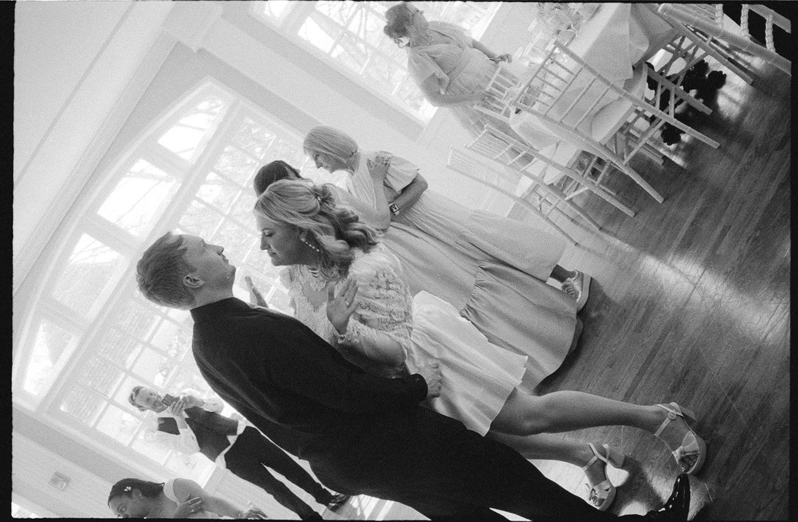 A black and white photo of a wedding ceremony, with the bride and groom facing each other and holding hands, surrounded by bridesmaids and a groomsman in a bright, elegant room with large windows and wooden floors.