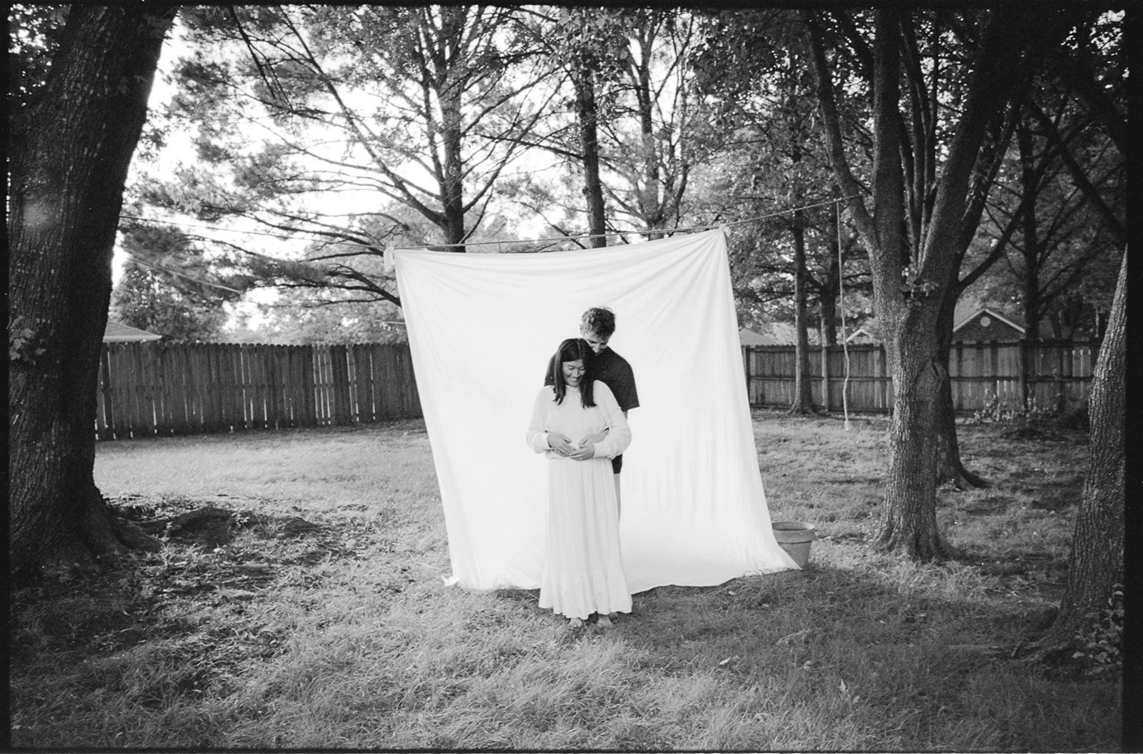 A couple standing outdoors in front of a white fabric backdrop, surrounded by trees and a wooden fence.