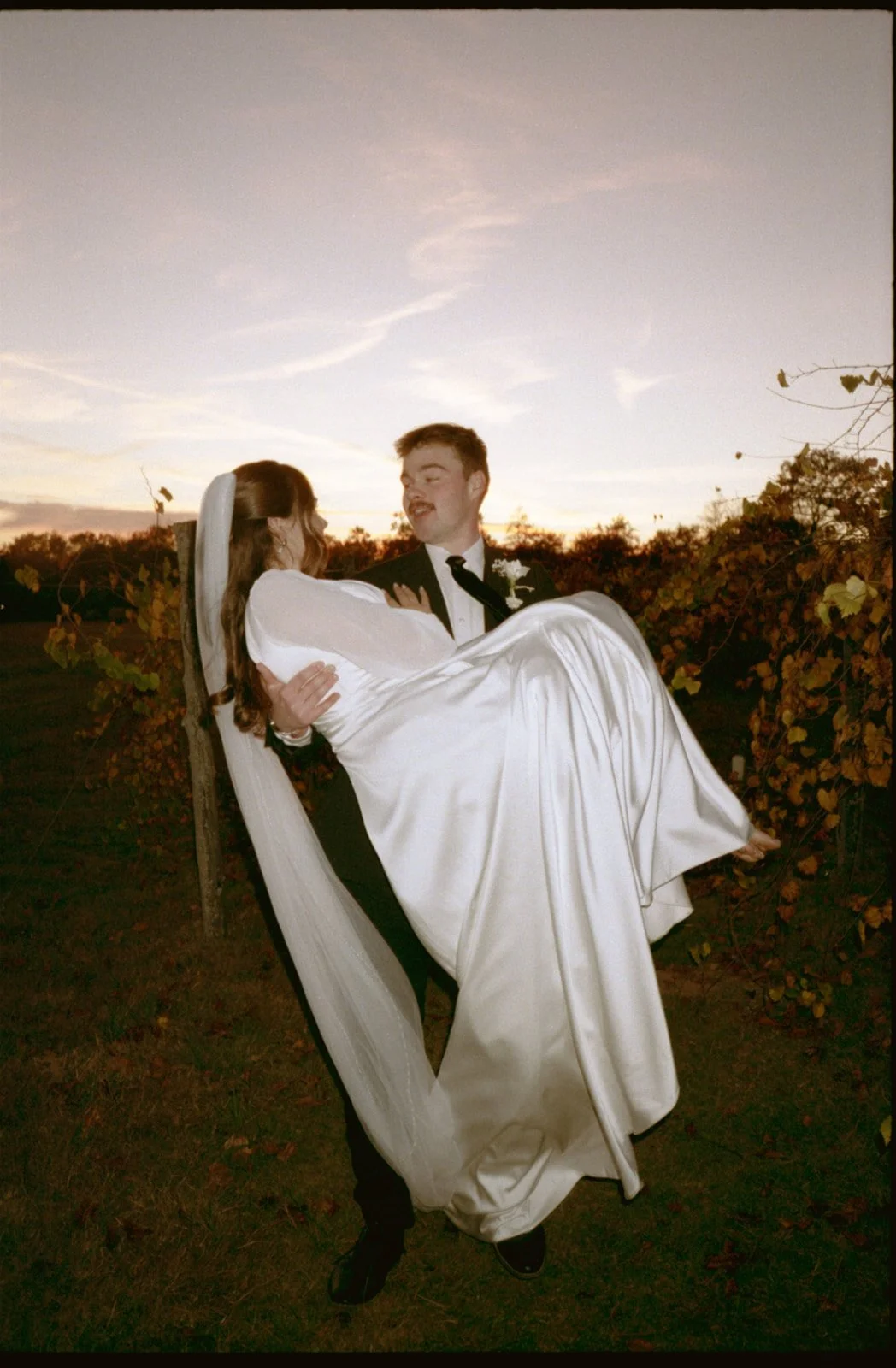 A groom, dressed in a tuxedo, holds a bride in a white wedding gown and veil, outdoors during sunset, with a backdrop of foliage and a colorful sky.
