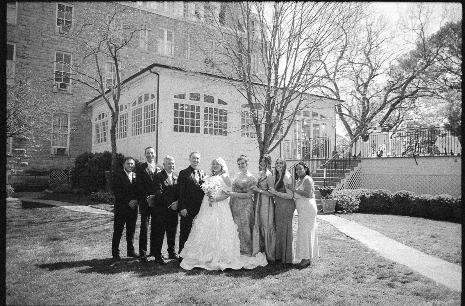 Group of people dressed in wedding attire standing on a lawn outside a building with trees and stairs.