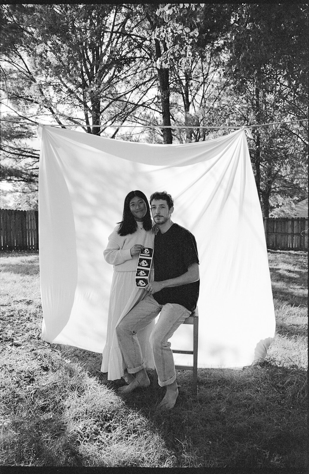 A black-and-white photo of a woman and a man posing outdoors in front of a fabric backdrop, with trees and a wooden fence in the background. The woman is standing, holding ultrasound images, and the man is sitting on a stool, holding the ultrasound p
