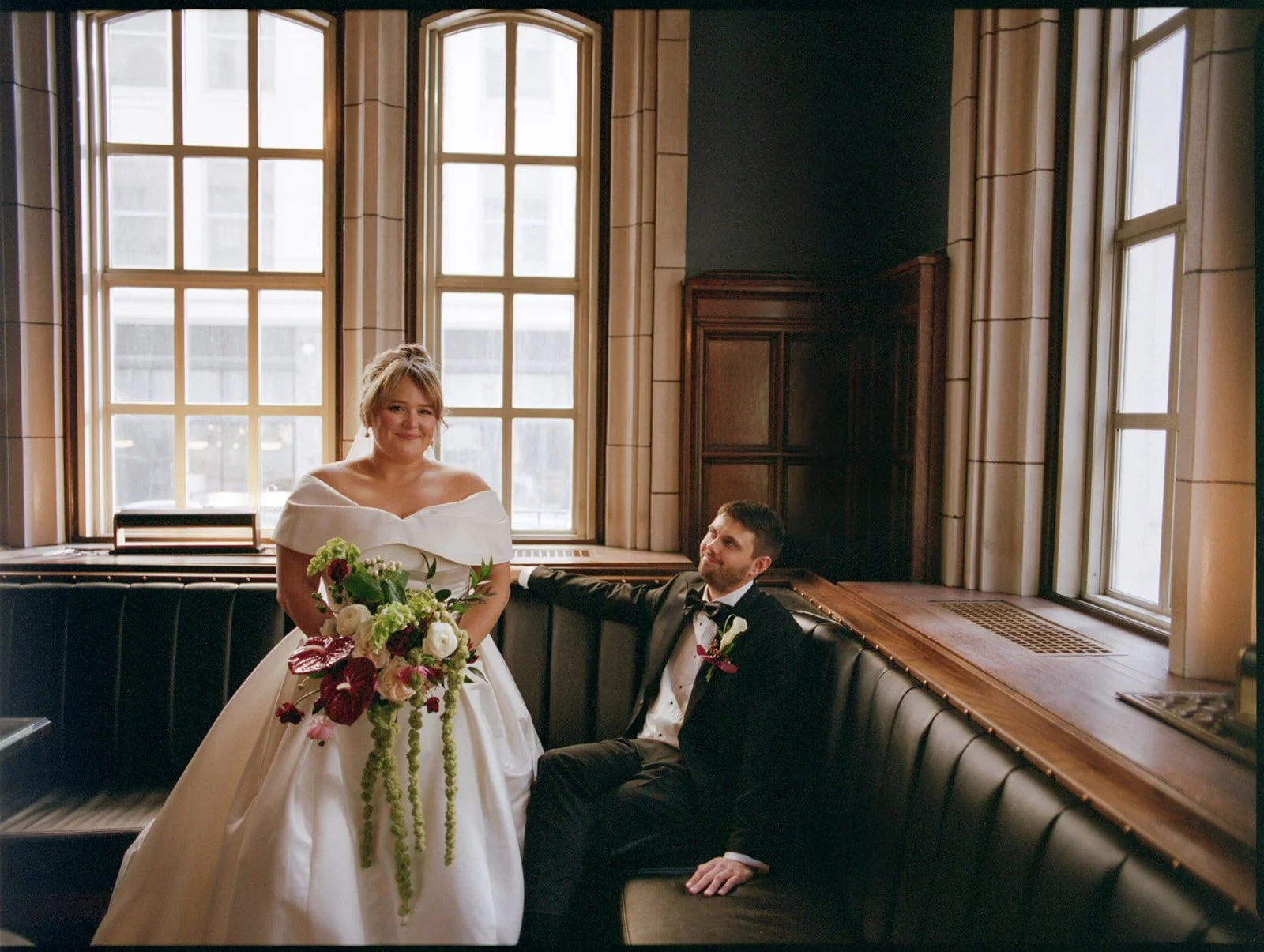 A bride and groom in wedding attire inside a room with large windows and dark wood paneling. The bride is standing holding a bouquet, and the groom is sitting on a black cushioned bench, looking at her.