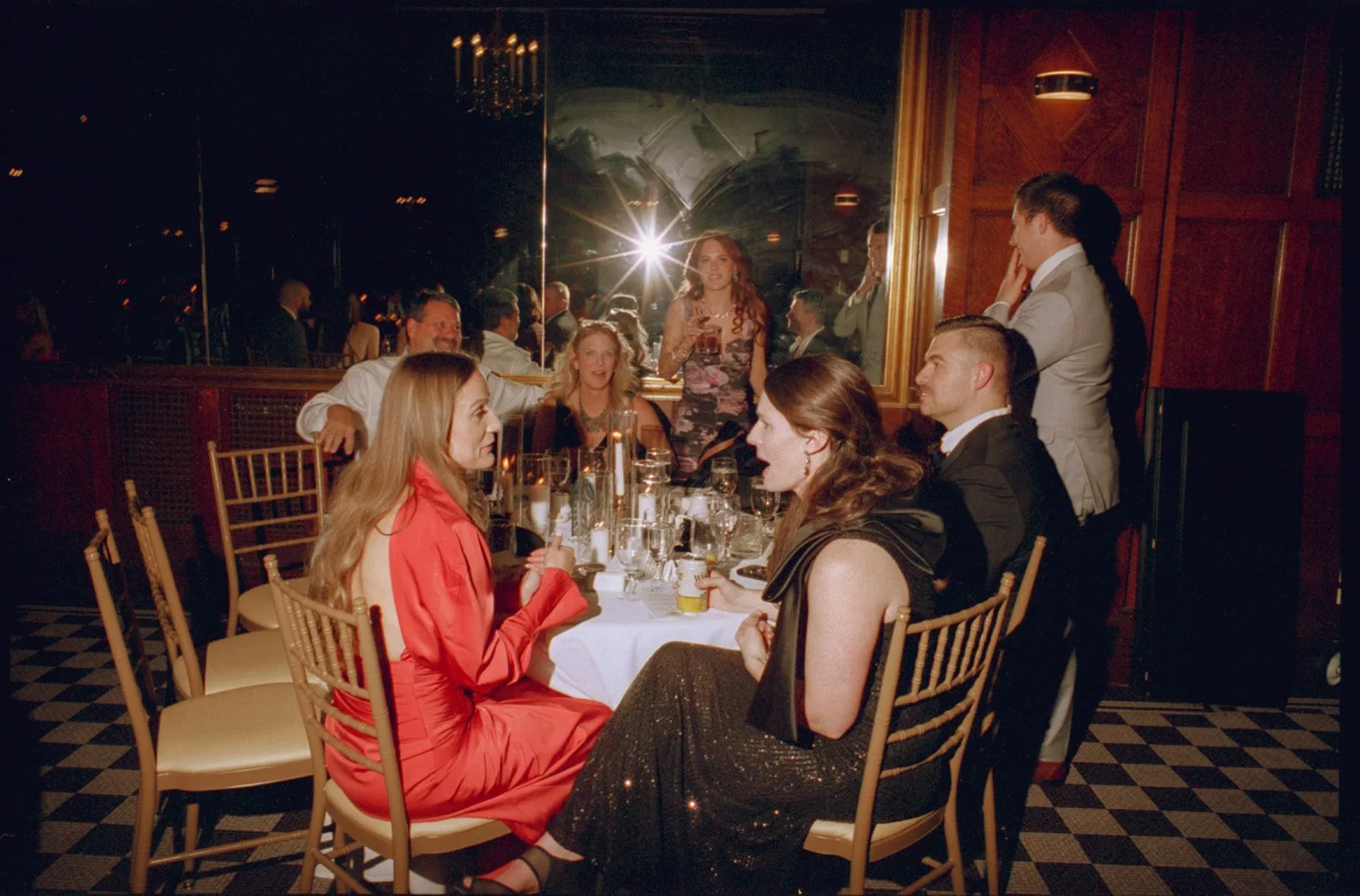 A group of guests at a formal dinner party in an elegant restaurant, with a mirror reflecting the scene and a chandelier hanging overhead.