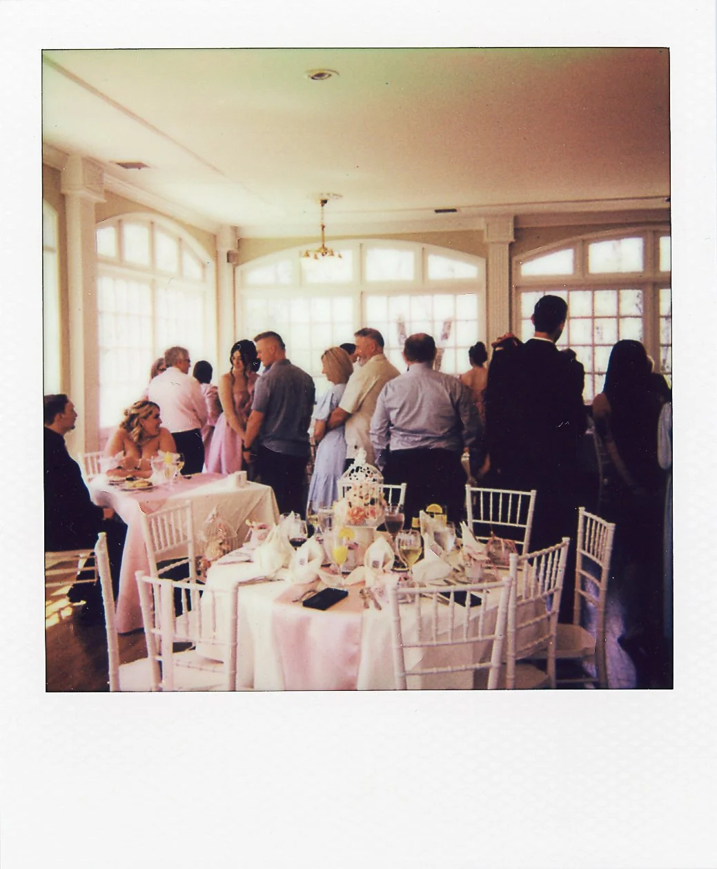 People gathered in a sunlit banquet room with round tables and white chairs, celebrating a special occasion, with some guests seated and others standing and talking.
