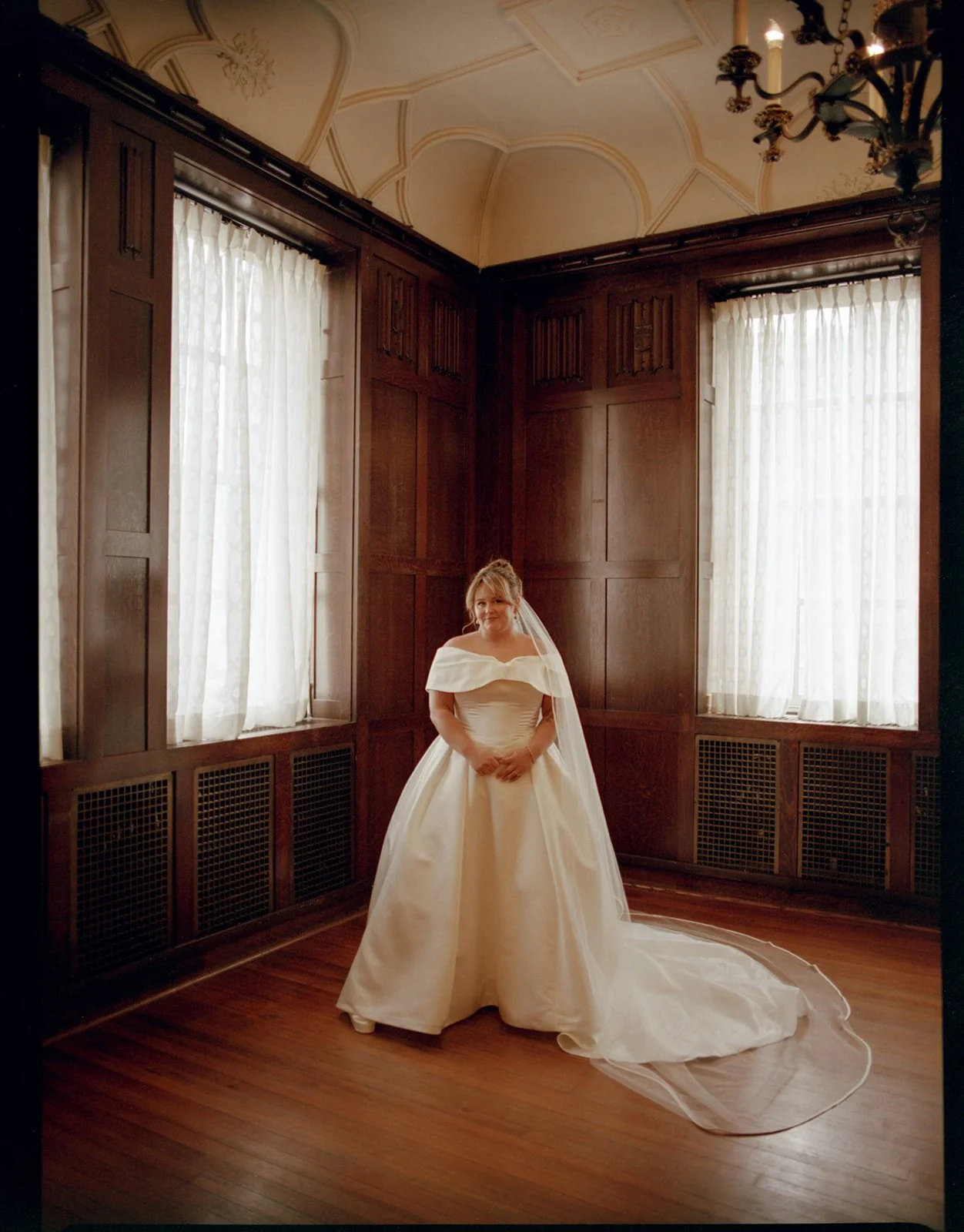A bride in a white wedding gown standing in a wood-paneled room with high ceilings and large windows with white curtains.