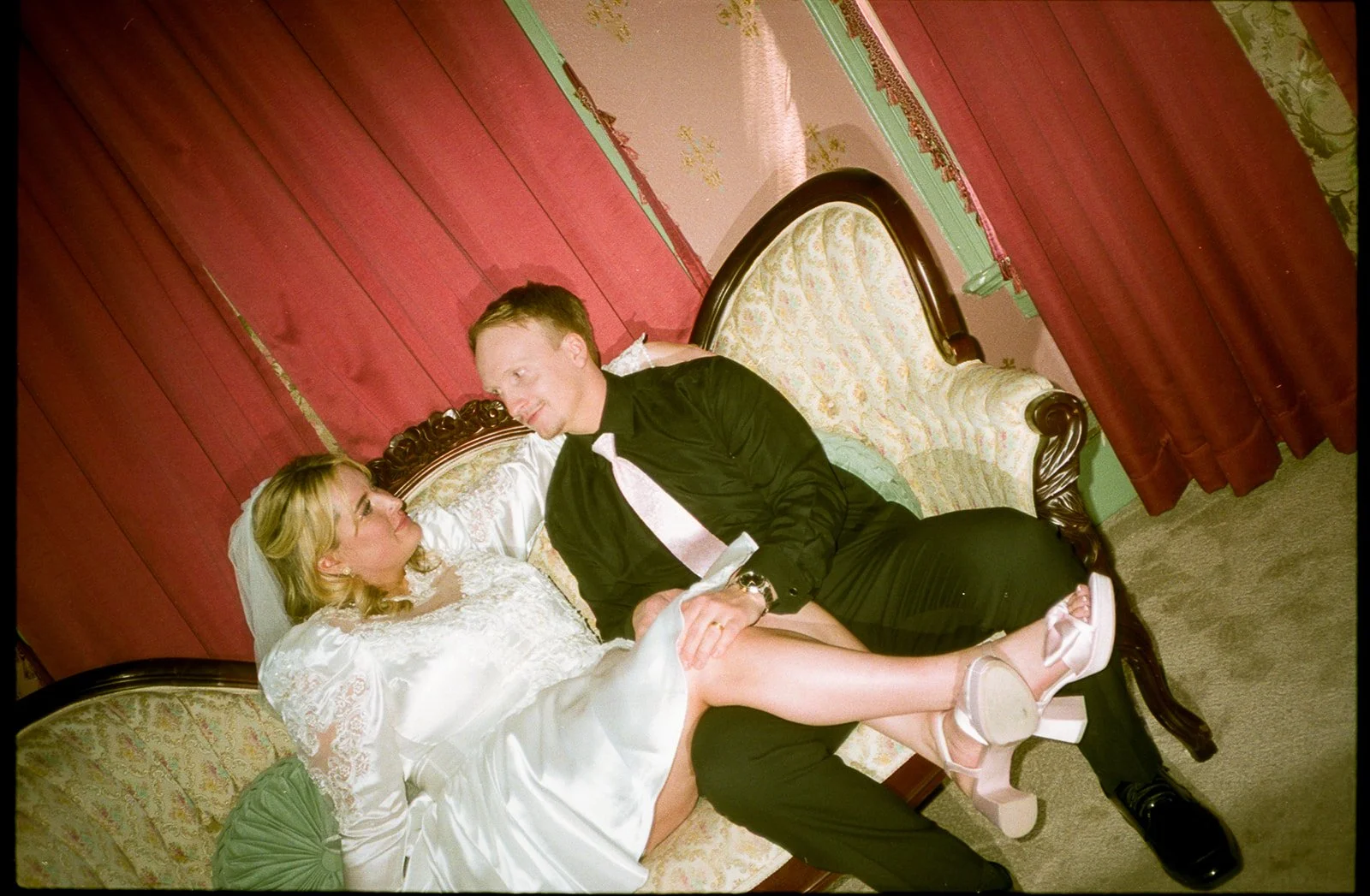 A bride and groom sitting on a vintage sofa, gazing into each other's eyes, in a decoratively decorated room with red curtains, green accents, and ornate furniture.