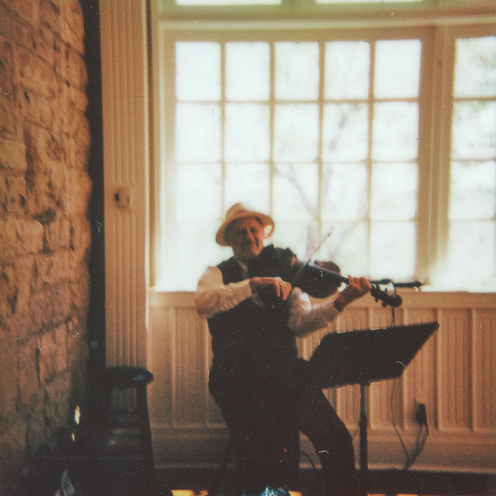 A person wearing a hat and vest playing the violin indoors near a large window.