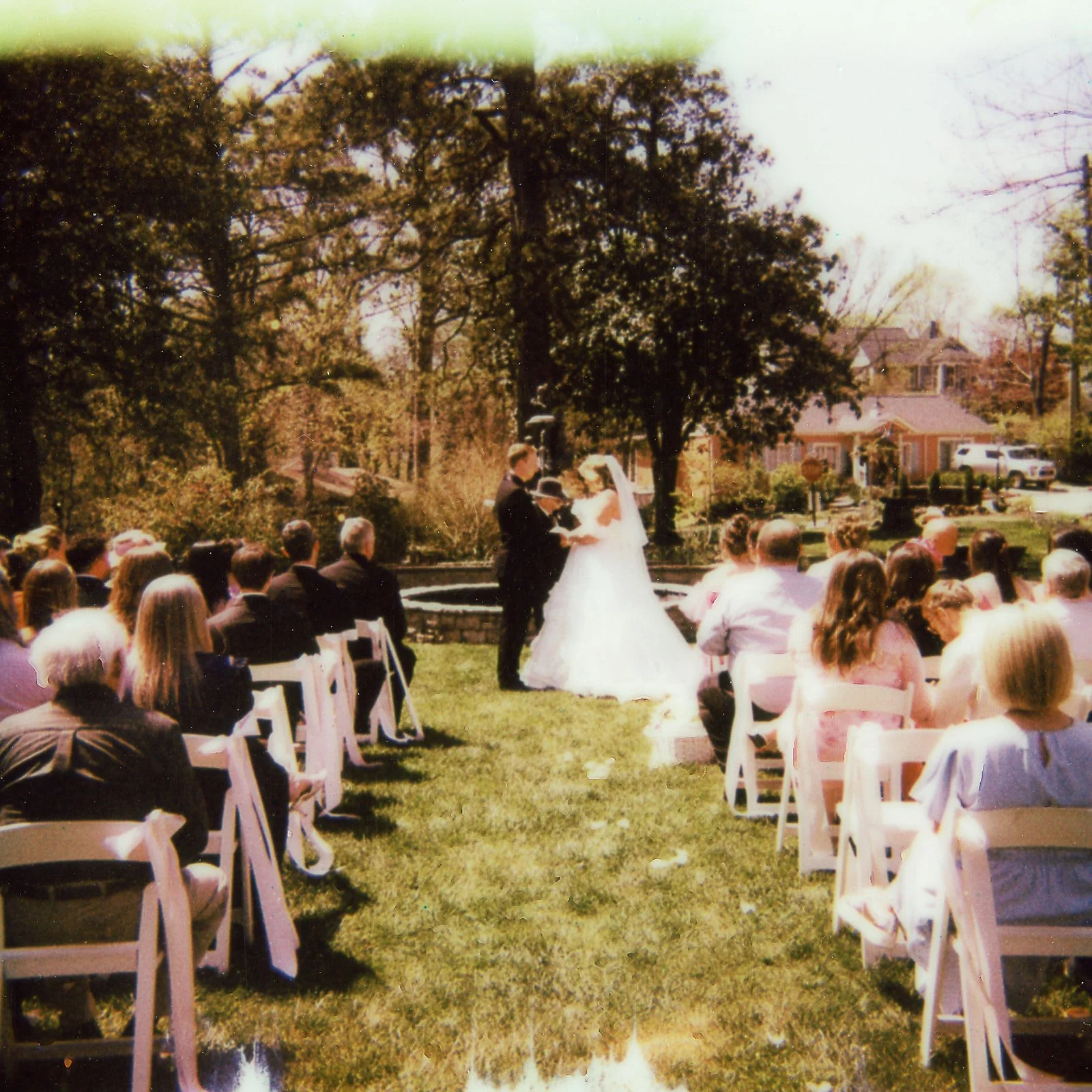 A wedding ceremony outdoors with a bride and groom exchanging vows in front of seated guests on a grassy area surrounded by trees.