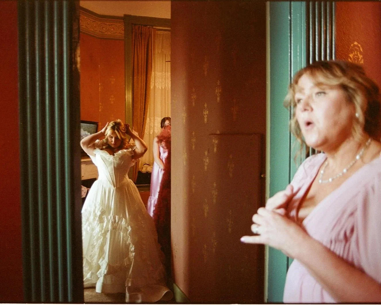 A woman in a wedding dress getting ready in front of a mirror, with another woman in pink observing her in a room decorated with patterned red wallpaper, gold curtains, and green door frames.