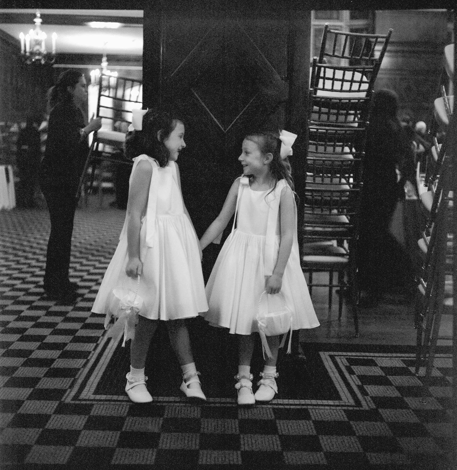 Two young girls in white dresses with bows in their hair, holding small baskets, standing on a checkered floor, smiling and holding hands, in a dimly lit room with stacked chairs and a dark wooden partition nearby.
