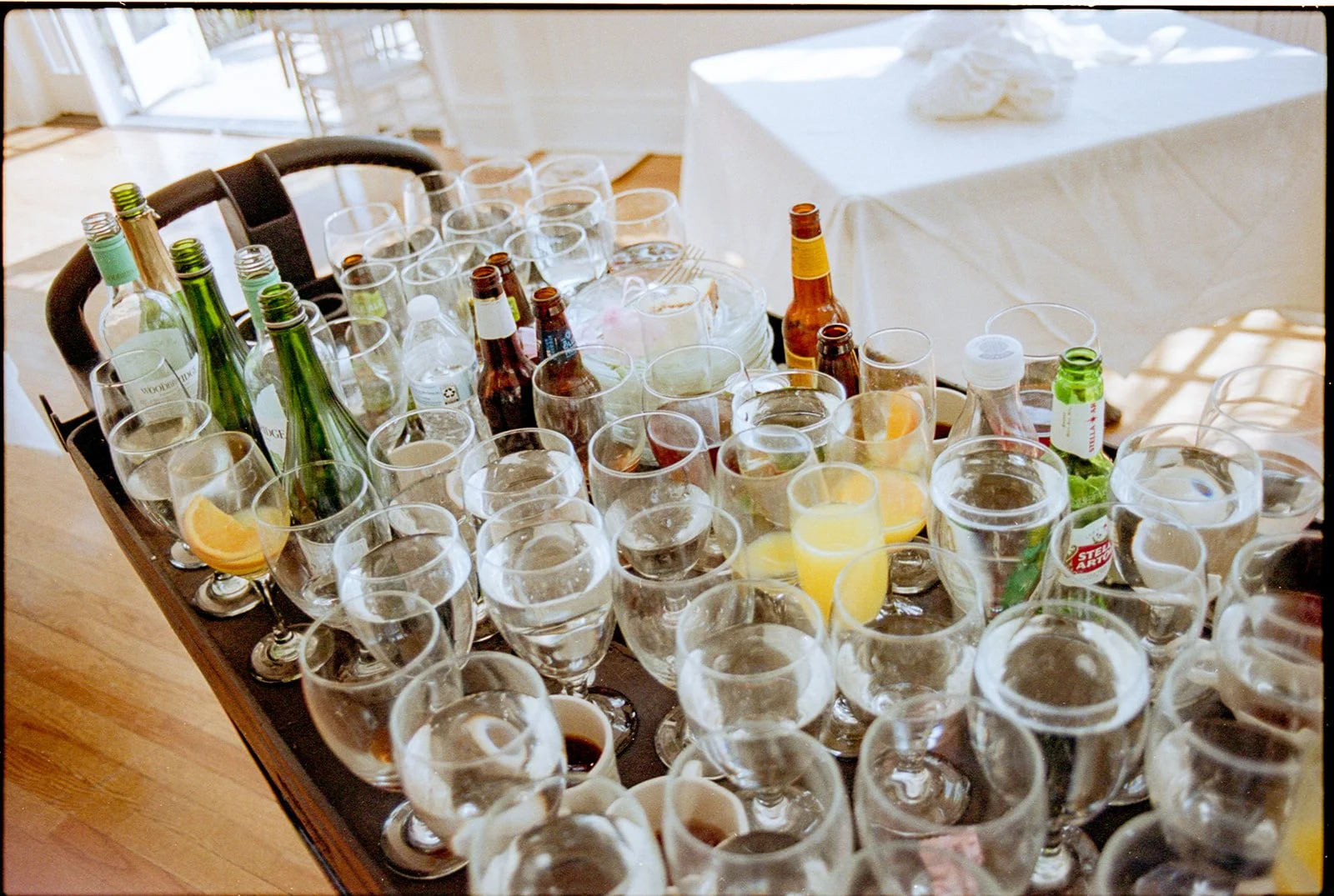 A table filled with various empty and half-filled glasses, bottles of soda and alcohol, and a few lemon wedges, set in a room with a wooden floor and large windows.