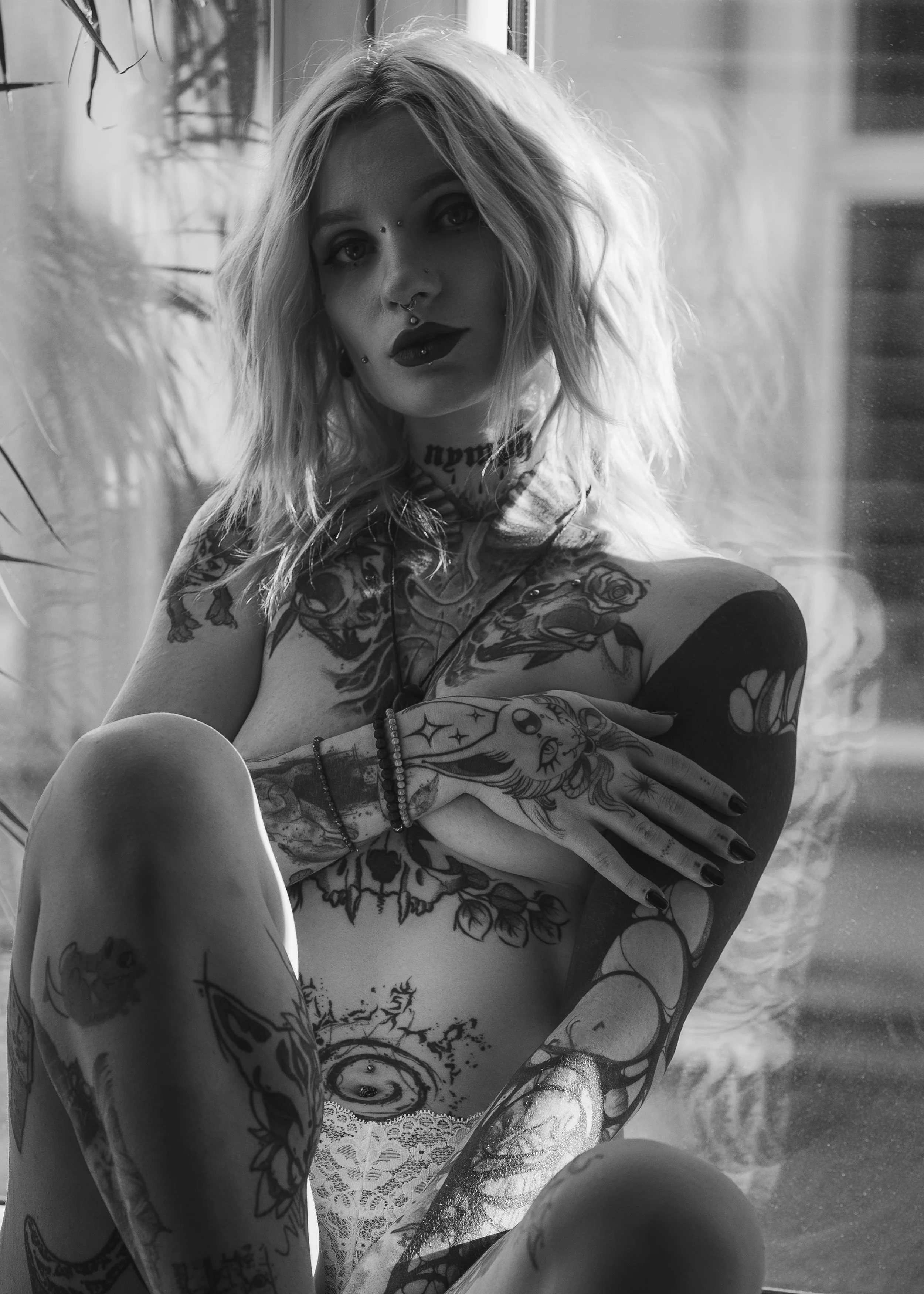 Schwarz-Weiß-Aktfotografie Gießen – Tätowierte Frau mit blondem Haar, dunklem Lippenstift, Piercings, am Fenster. Charakterstarkes, künstlerisches Frauenportrait mit urbaner Note.