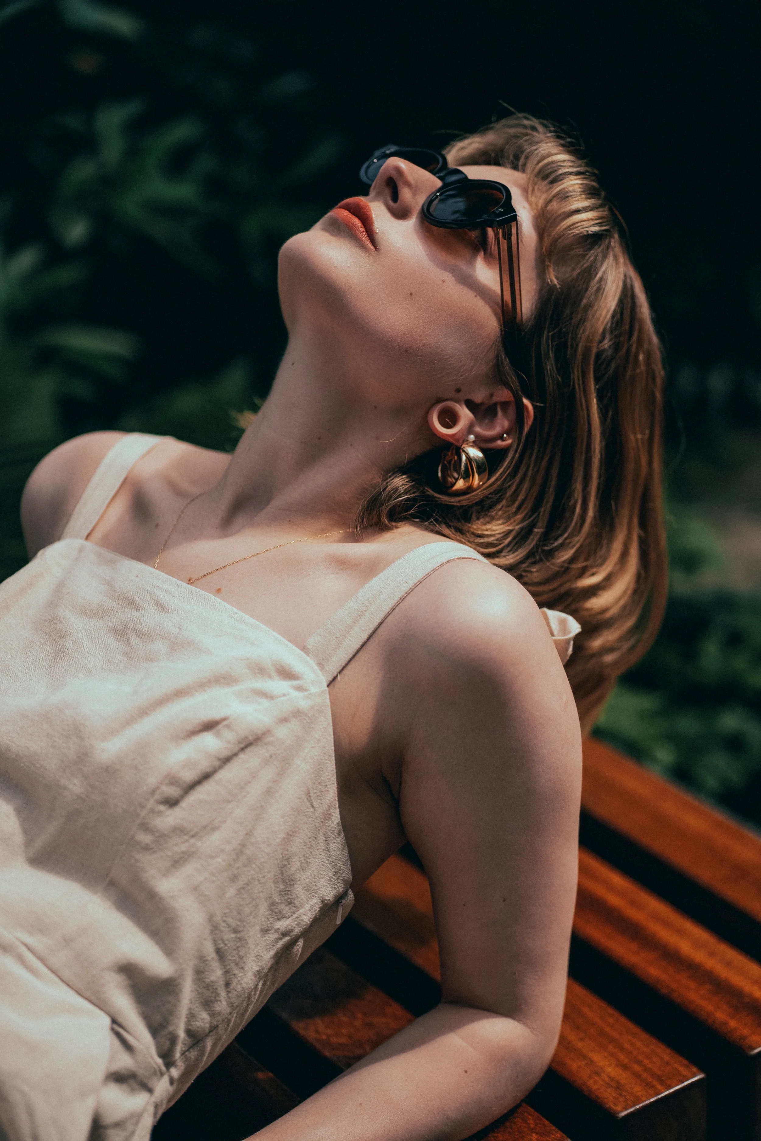 Lifestyle Portrait Wiesbaden – Frau mit Sonnenbrille, goldenen Ohrringen, liegt entspannt auf Holzbank im Grünen. Modernes Frauenportrait, entspannte Atmosphäre, modischer Look.