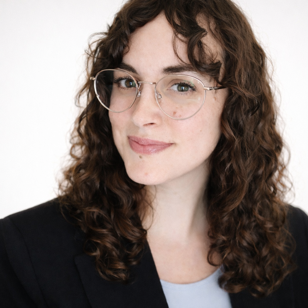 A woman with long, curly brown hair, wearing glasses, a black blazer, with a white background.