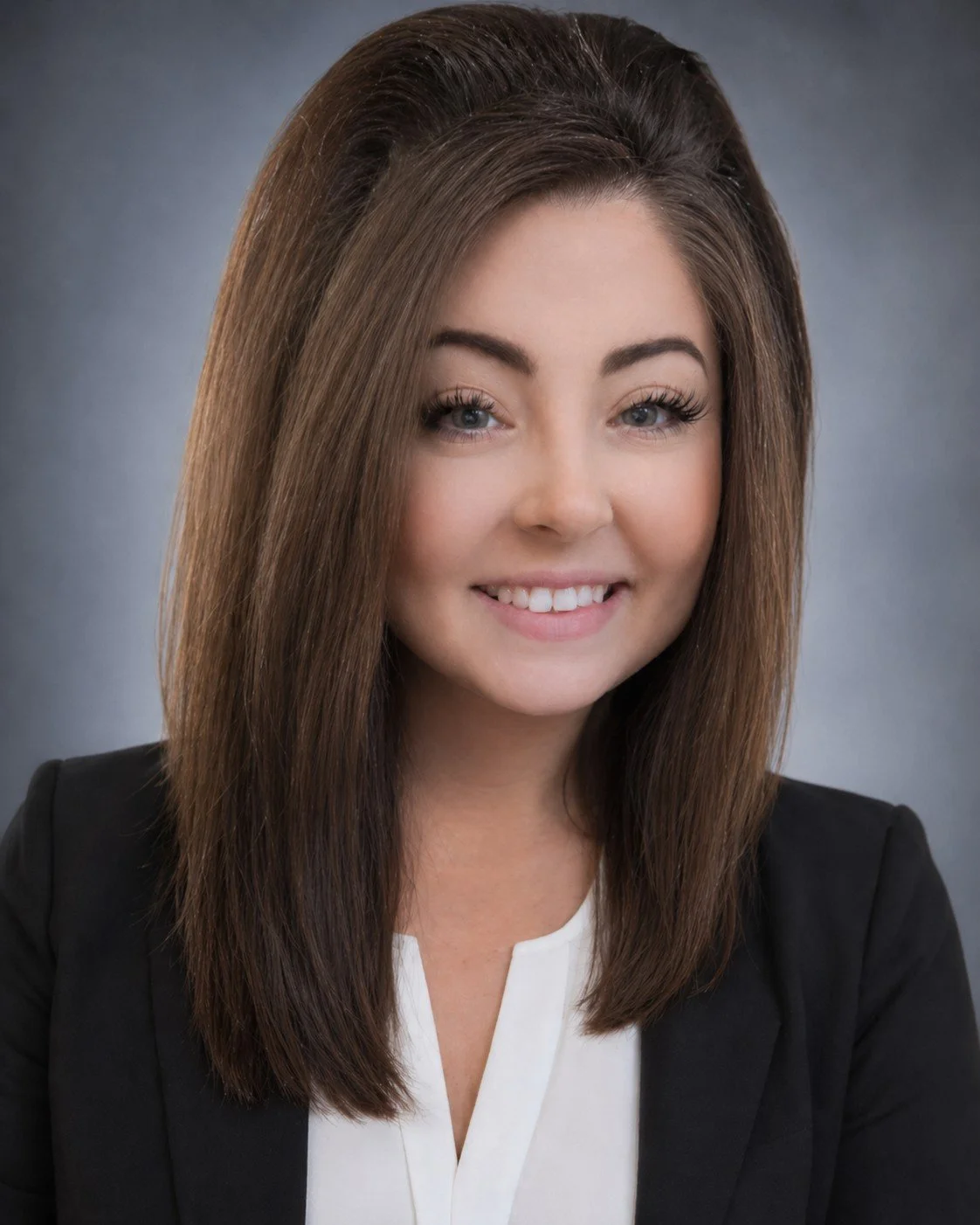 A professional headshot of a woman with straight brown hair, smiling, wearing a black blazer over a white top, against a blurred gray background.
