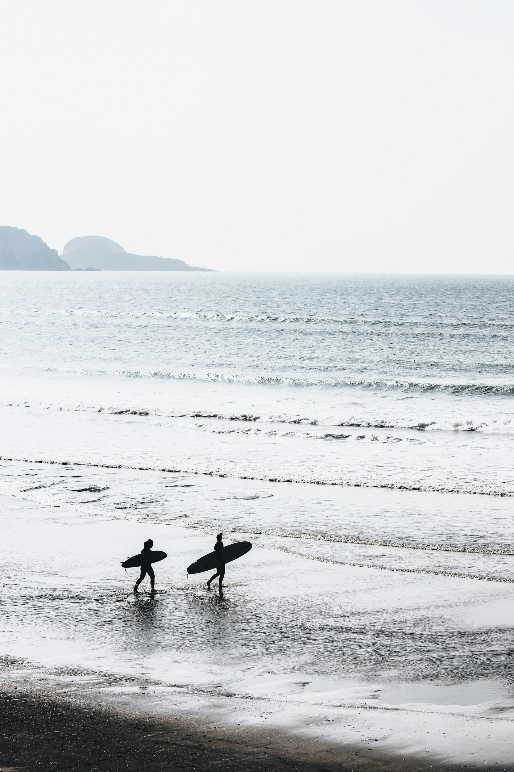 Silhouettes of two surfers walking on the beach with surfboards, ocean waves, and distant cliffs in a monochrome tone.