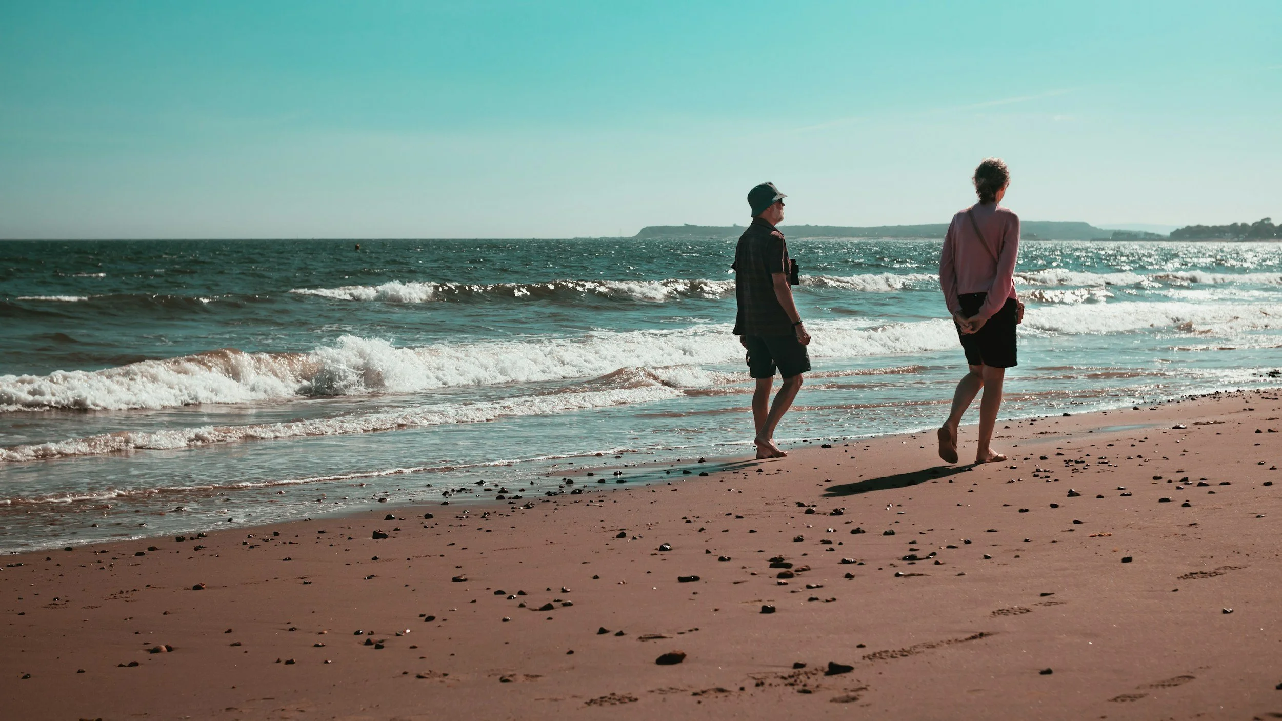 Two people walking along a sandy beach with small rocks, waves crashing, and a distant landmass in the background during daytime.