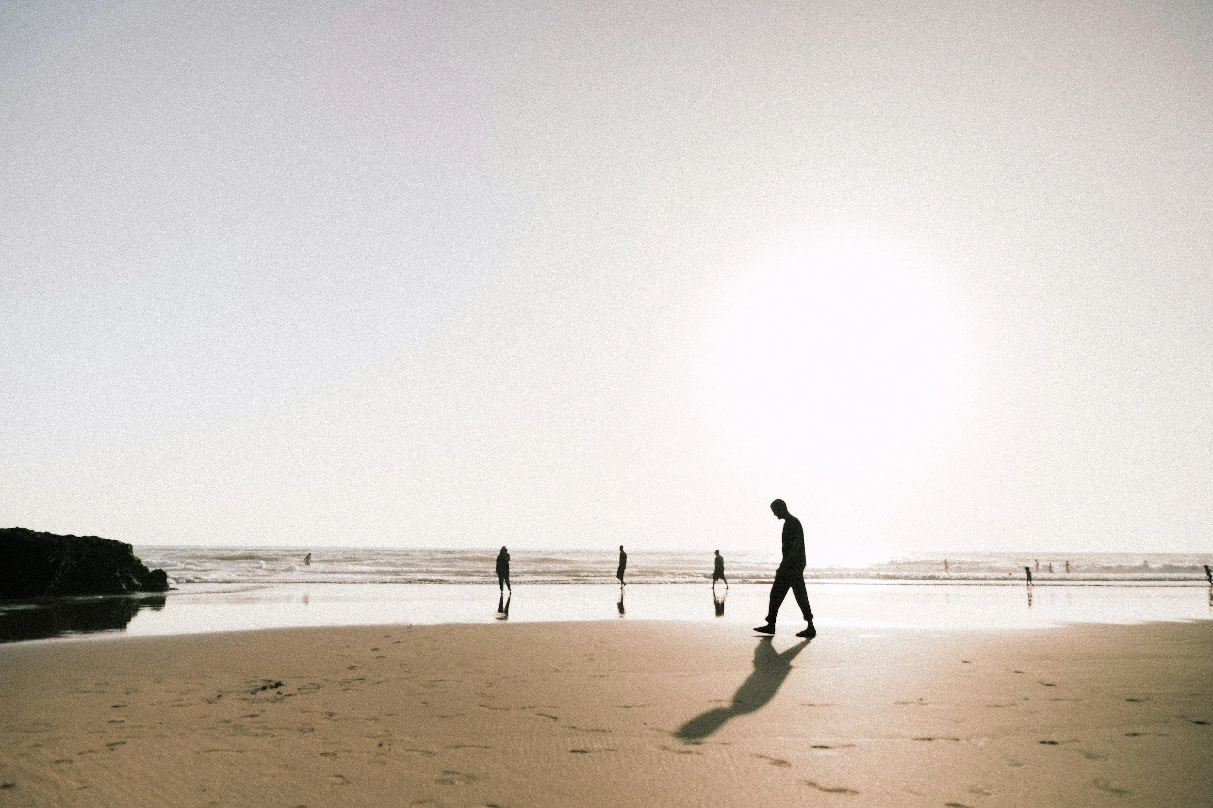 Silhouettes of people walking on a beach during sunset or sunrise, with the ocean and a cloudy sky in the background.