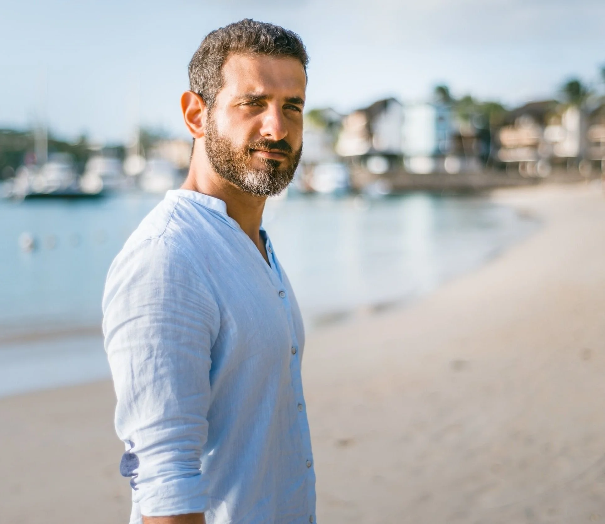 A man with a beard and short hair wearing a white shirt standing on a beach with boats and houses in the background wondering what the point of his life is.