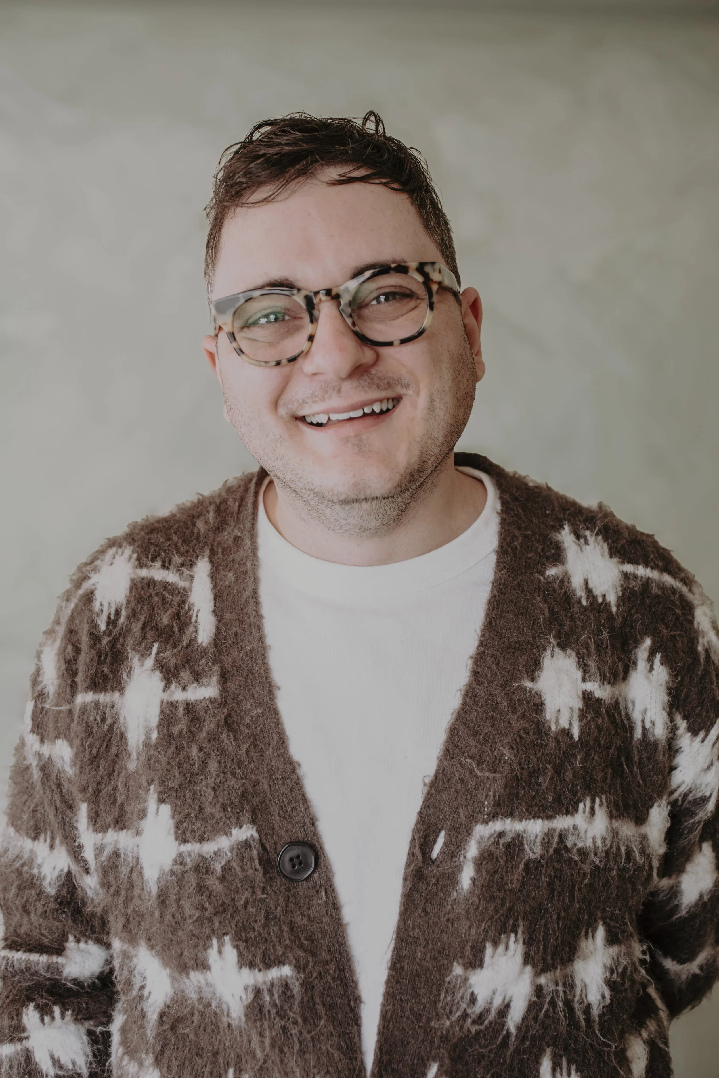 A smiling man wearing tortoiseshell glasses and a brown and white patterned fuzzy cardigan.