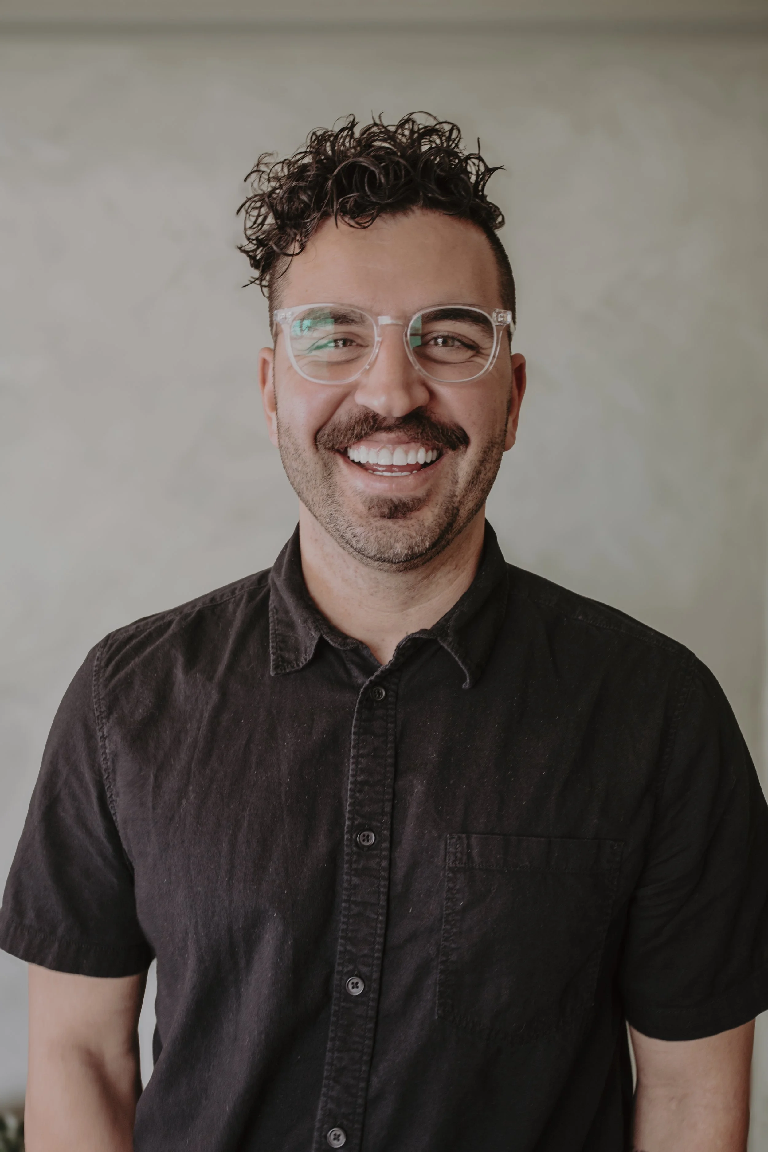 A young man with curly hair, glasses, and a beard, wearing a black shirt, smiling he brightens the photo.