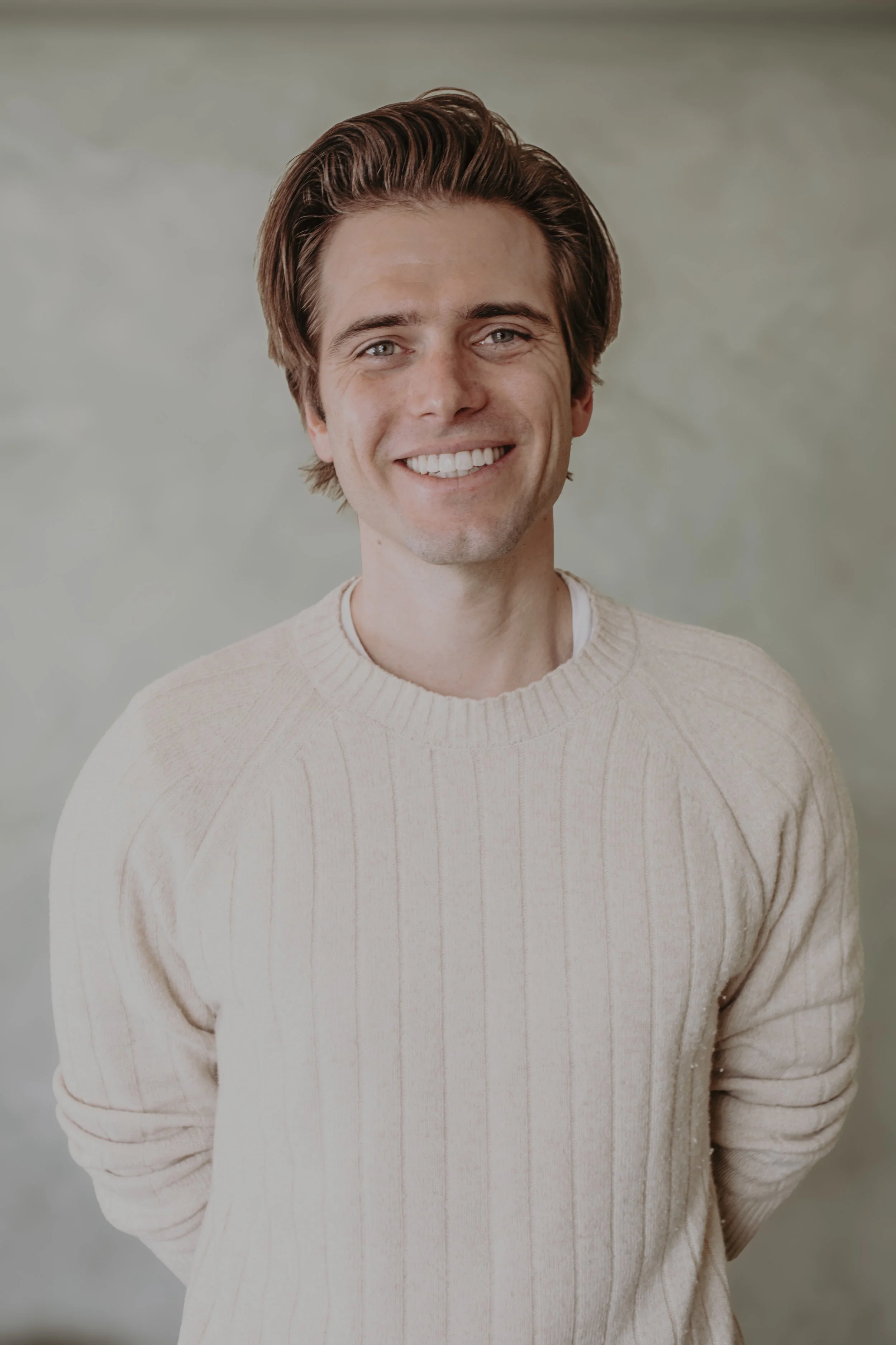 A young man with light brown hair, smiling, wearing a cream-colored sweater, against a neutral background.