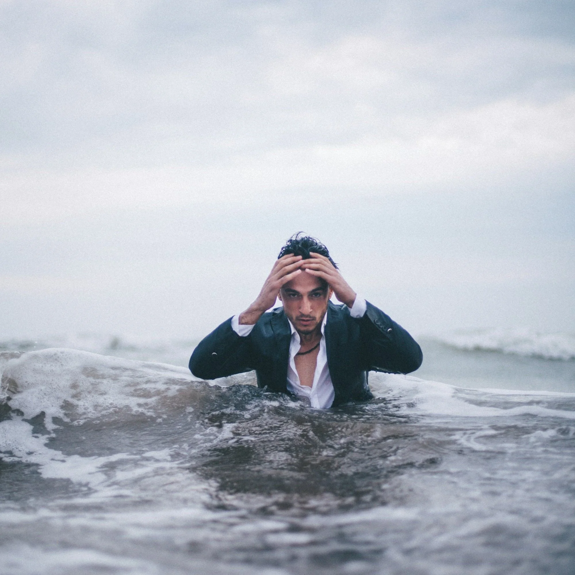 A man in a wet suit holding his head in the ocean, with a cloudy sky overhead.