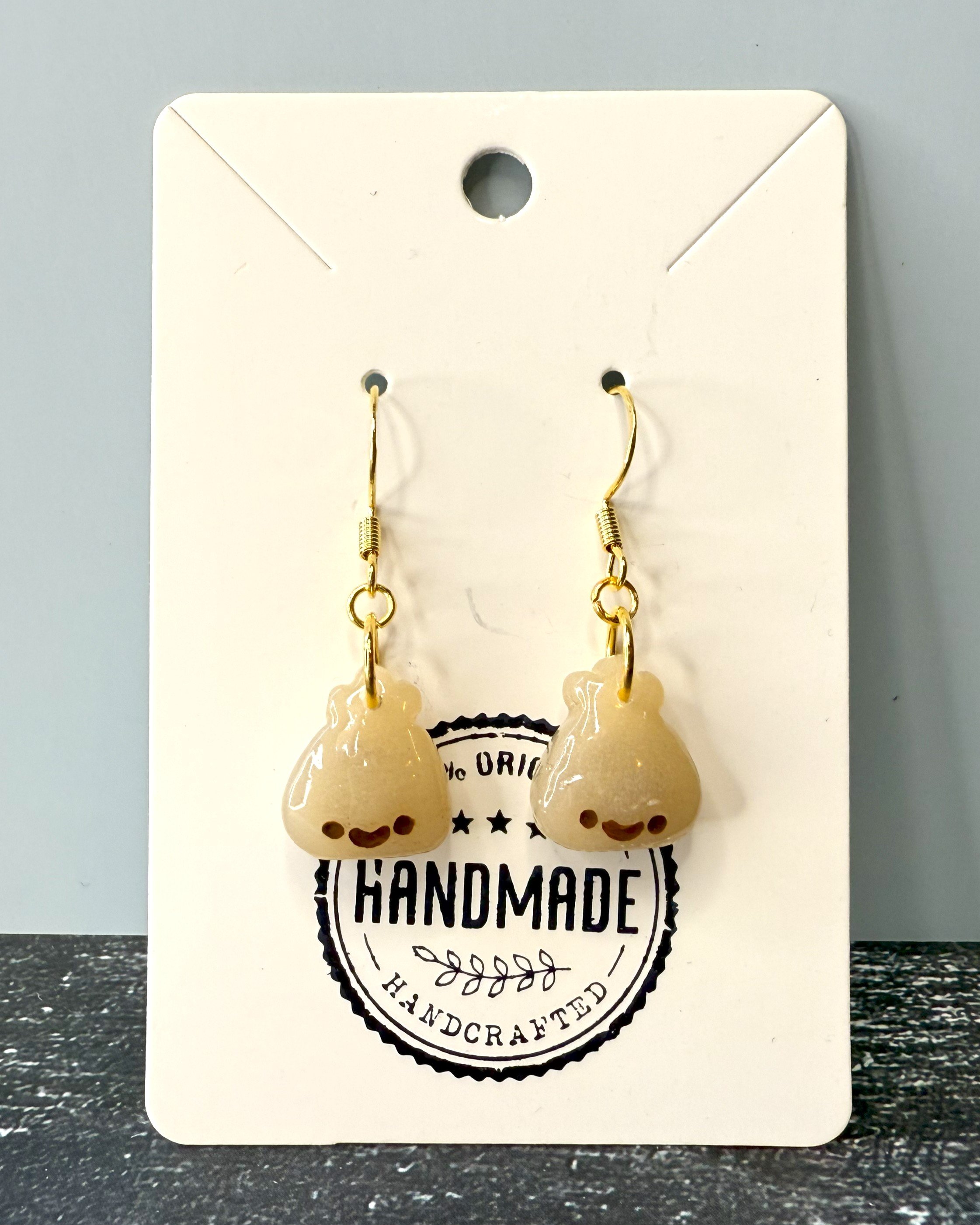 Pair of handmade polymer clay earrings shaped like smiling dumplings, attached to a branded display card and photographed on a clean, neutral background
