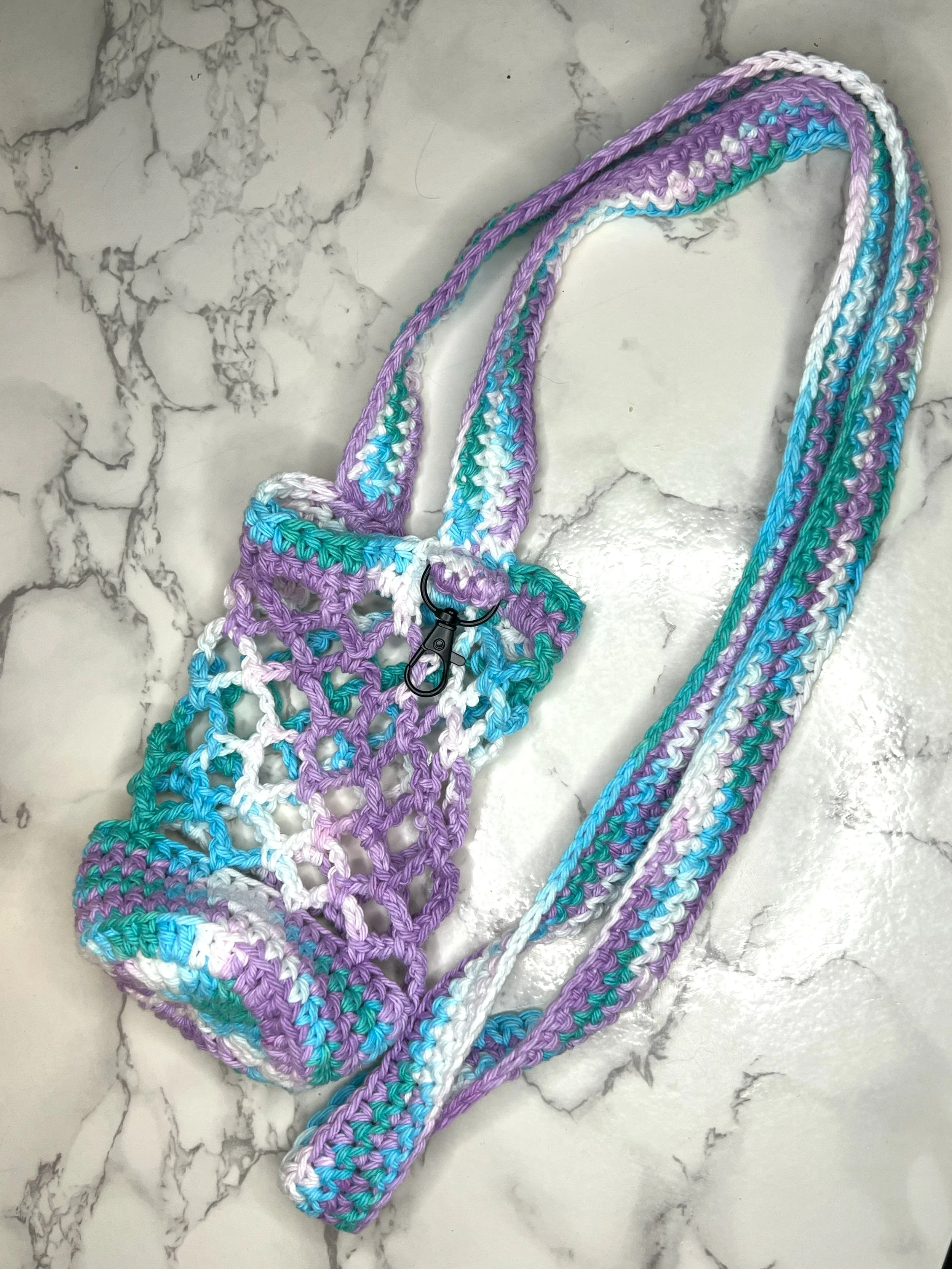 Crochet water bottle holder in blue, purple, and white netting with a keychain clip, laid flat on a marble tabletop