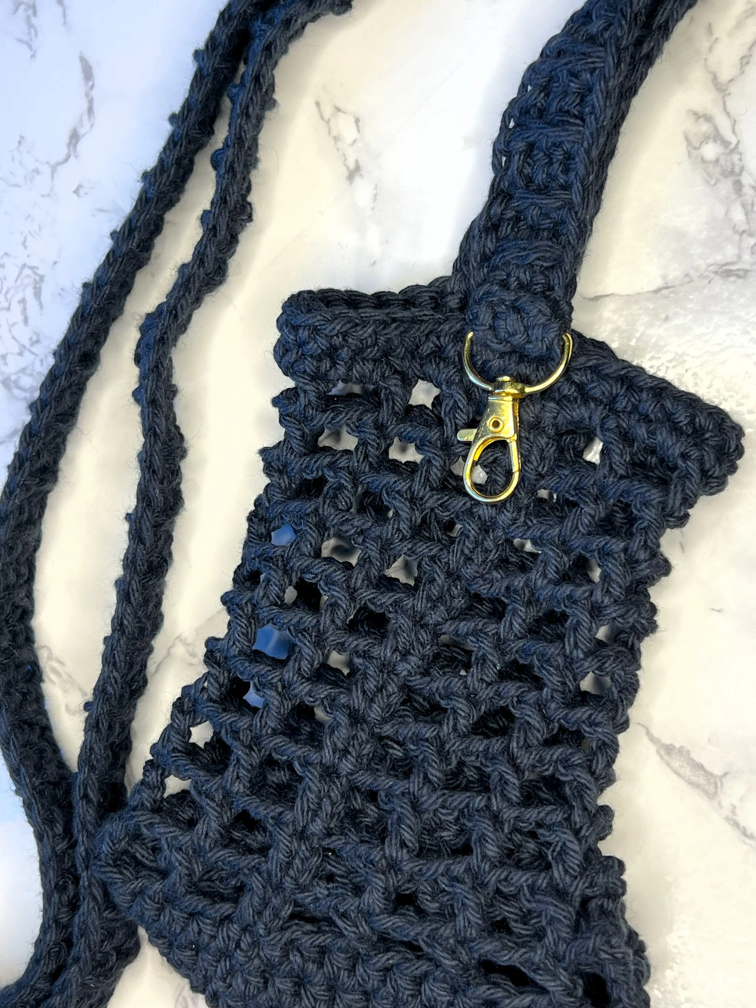 Black crochet water bottle carrier in a square net design with a keychain clip, shown laying flat on a white and black marble background