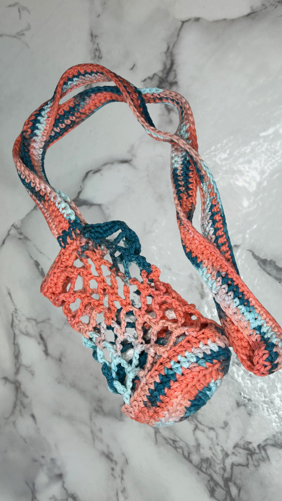 Handmade crochet water bottle holder in a pink and blue net pattern, laid flat on a white and black marble tabletop