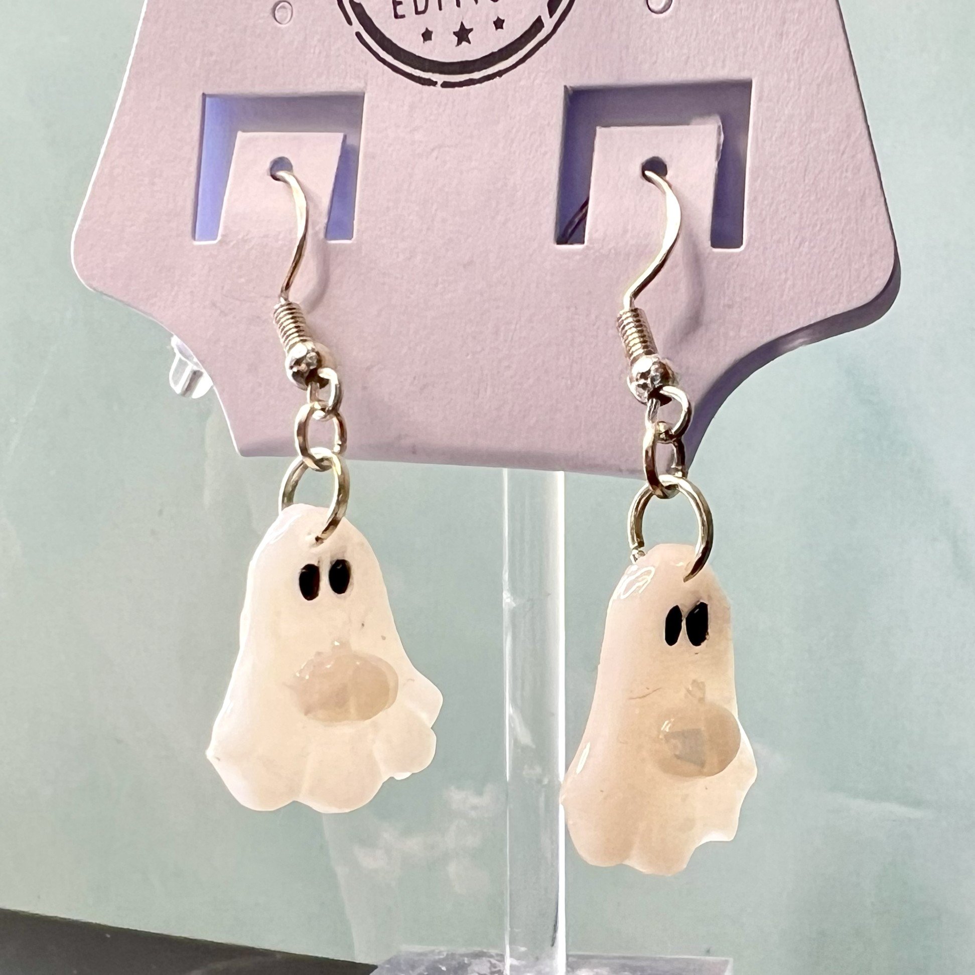 Translucent ghost earrings with iridescent opal pumpkins shown on a branded display card resting on a black and white marble tabletop