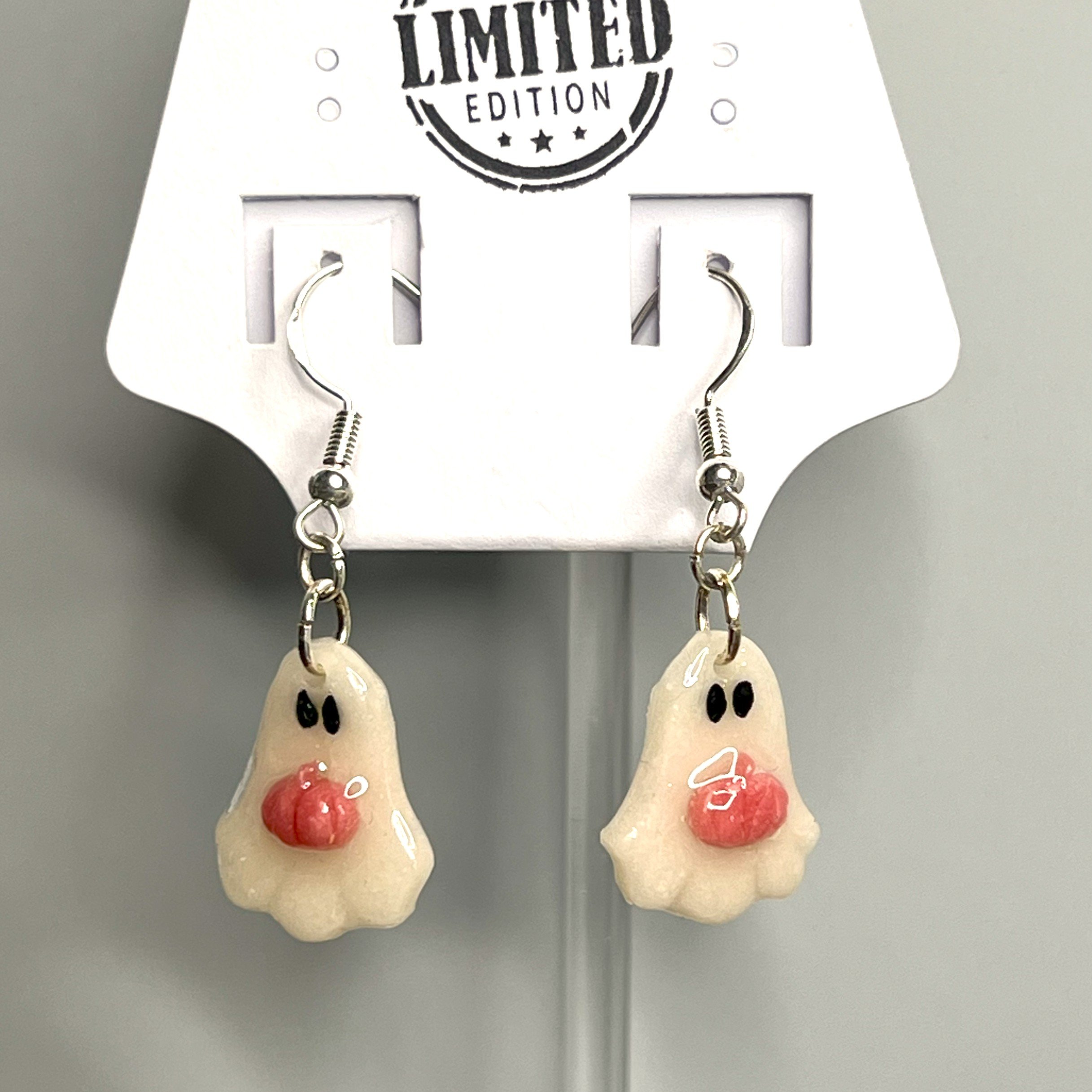 Ghost earrings made from translucent clay with a thermochromic pumpkin that changes color, shown on a display card atop a marble tabletop