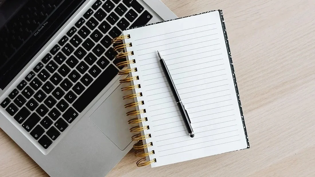 Open notebook and pen next to a laptop on a desk.