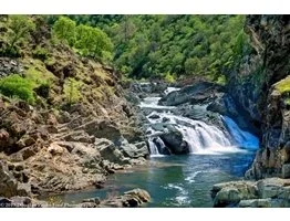 A river with a small waterfall flowing through a rocky landscape surrounded by green trees.