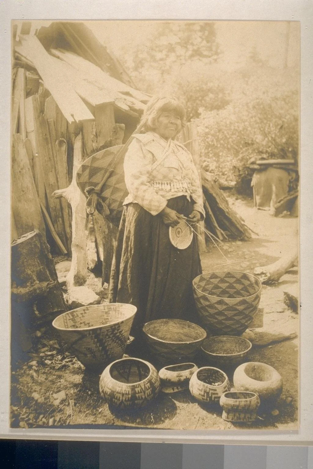An indigenous woman standing outdoors near a rustic wooden structure, surrounded by woven baskets with geometric patterns, holding a small object with strings.