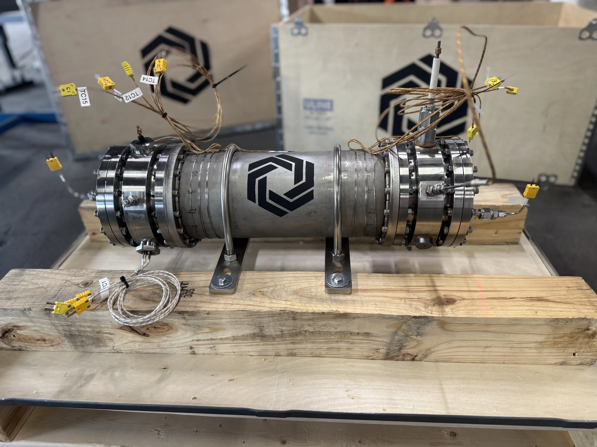 A large metallic cylindrical device with multiple connectors and wires, resting on a wooden platform in an industrial setting.