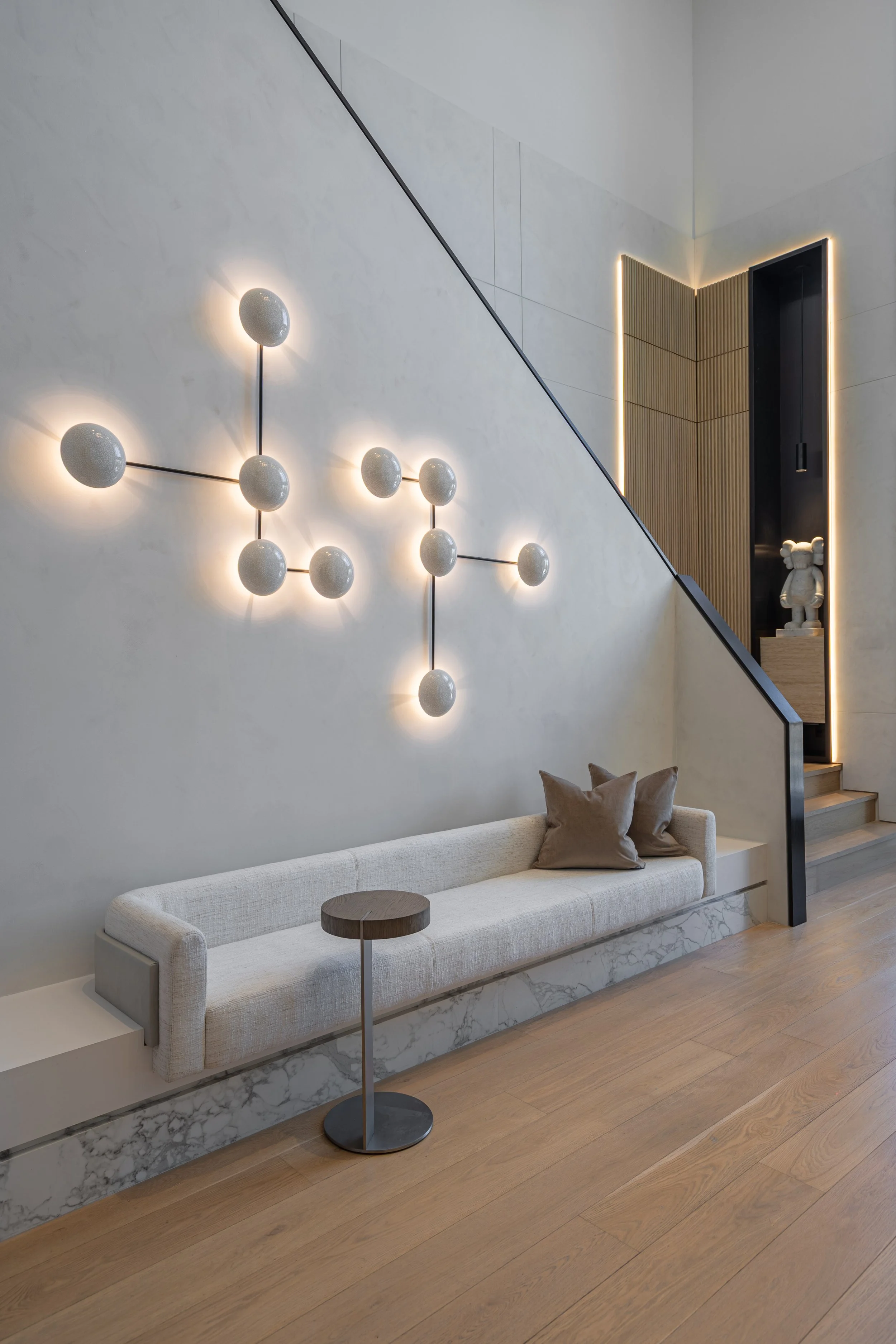 Modern living room with a long beige sofa, two brown pillows, a small round side table, a geometric wall light, and a staircase with a decorative sculpture at the top, featuring minimalist and neutral decor.