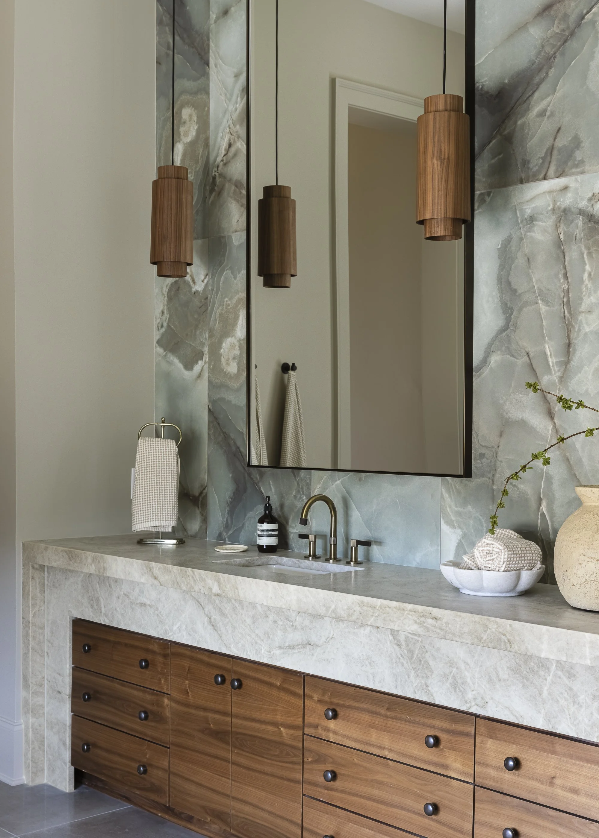 Modern bathroom vanity with marble countertop, wooden cabinets, large mirror with black frame, above-counter sink, black faucet, wooden pendant lights, decorative items including a beige vase, a small bowl with rolled towels, and a bottle of hand soap.