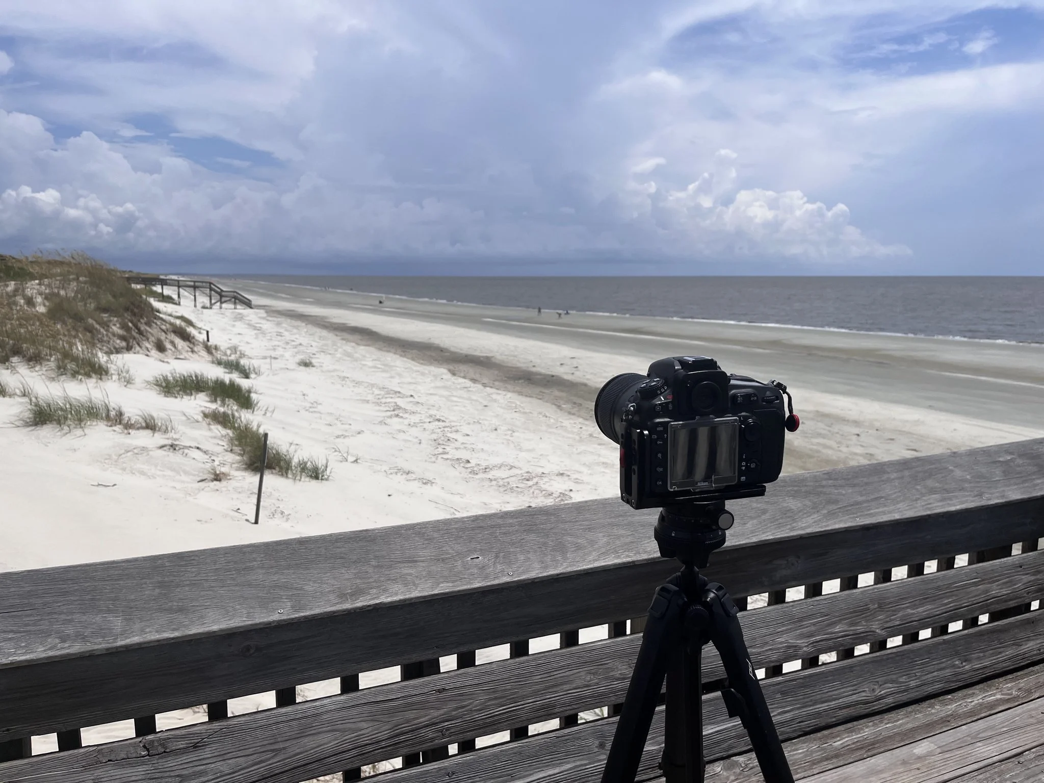 Camera on tripod facing a sandy beach with grass dunes, wooden staircase in the distance, and cloudy sky over the ocean.
