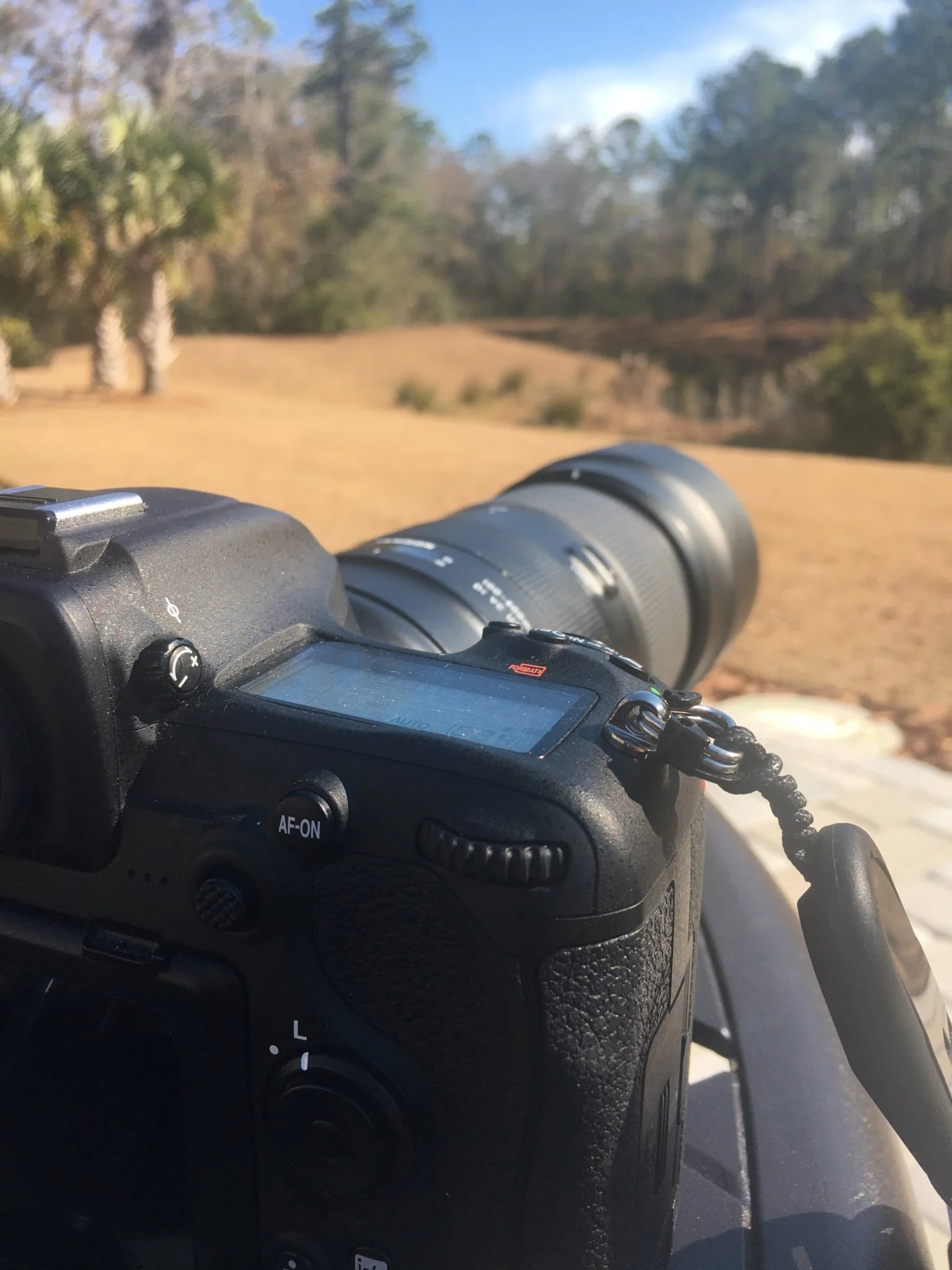 Close-up of a digital camera with a long lens, set outdoors near a body of water with trees and clear sky in the background.